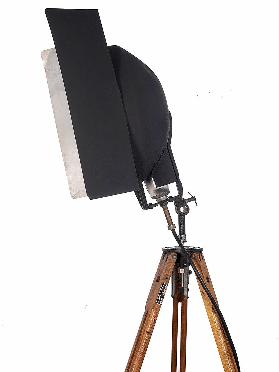 This is a wonderful looking industrial floor lamp. The original aluminum photographers flood light still has the articulating barn doors. The lamp is jointed so it tilts. The tripod is good one with that can be set to different heights. The wood