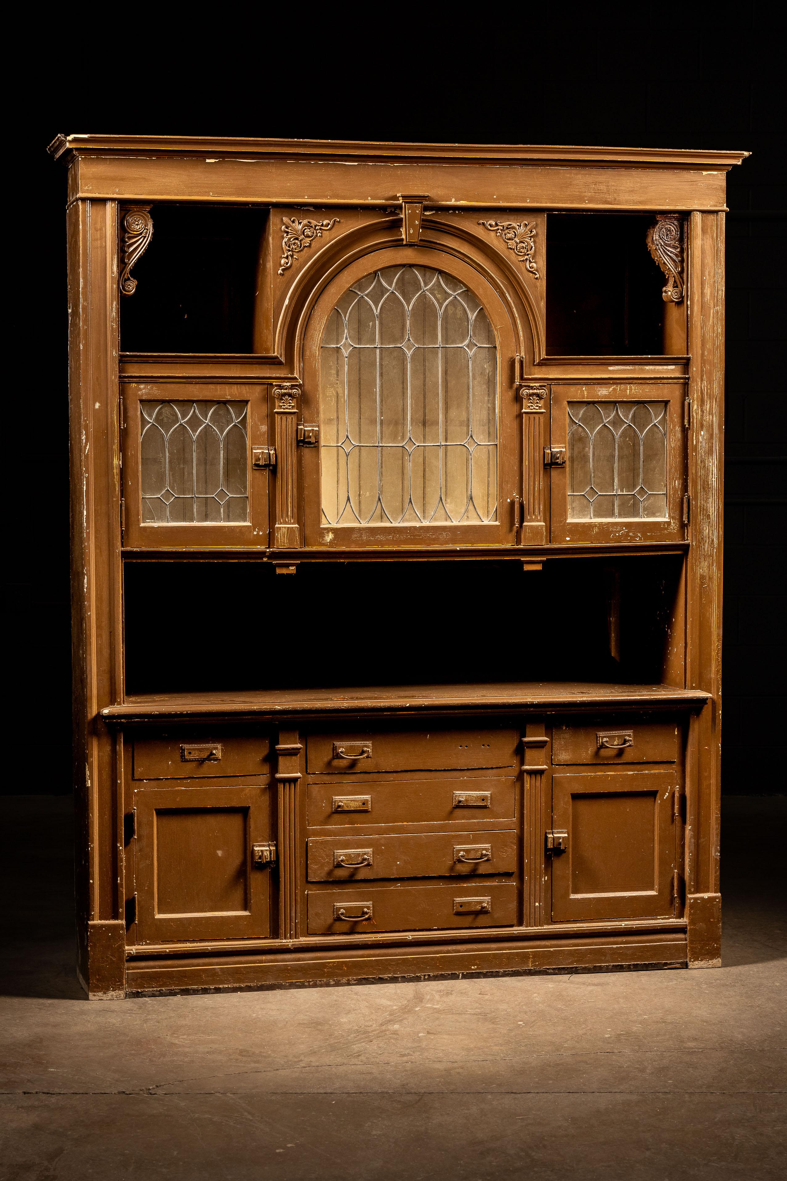 Antique wooden hutch with ornate carved embellishments.

This piece features a slightly chippy coat of light brown paint across its wooden exterior. There is a large central arched windowed cabinet flocked on either side by square cabinets of the