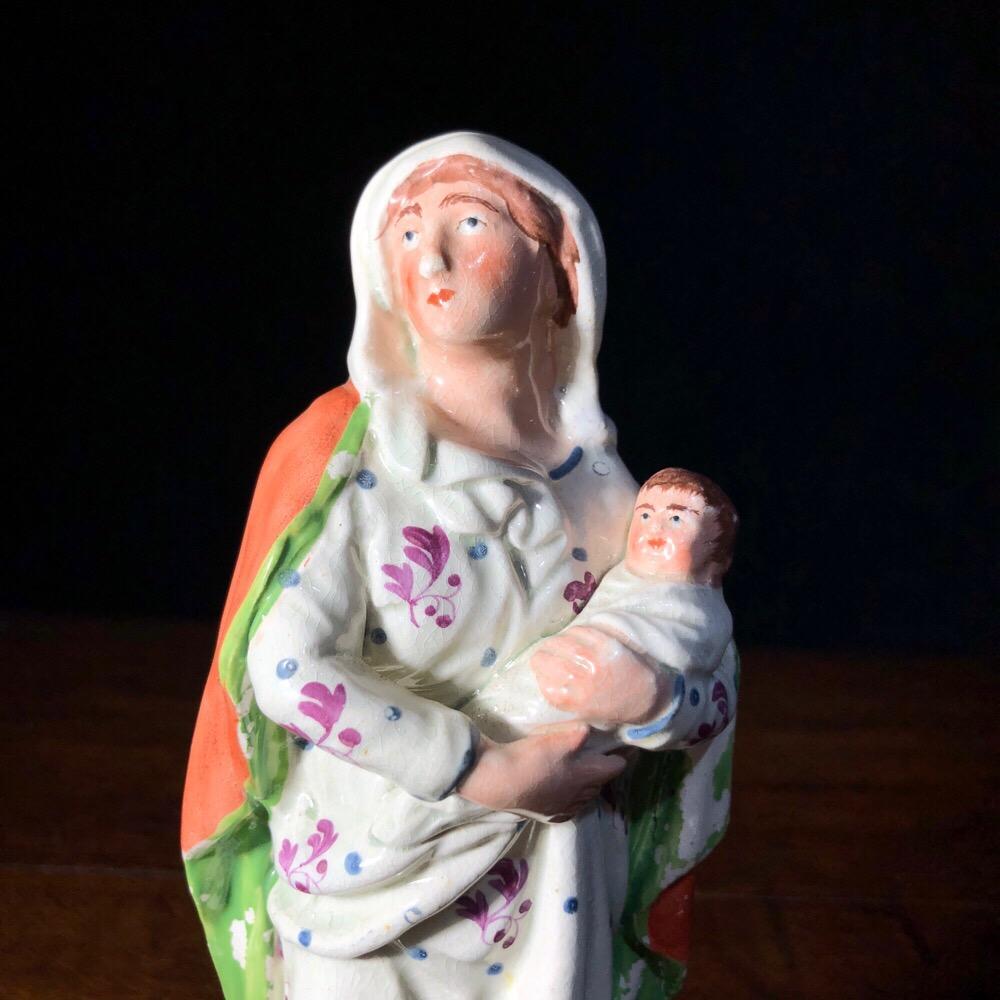 Early Staffordshire figure of ‘Virgin Mary’, modelled standing with child in her arms, on a low grassy mound set on a square plinth bearing the inscription ‘Virgin Mary’,
circa 1810.

 