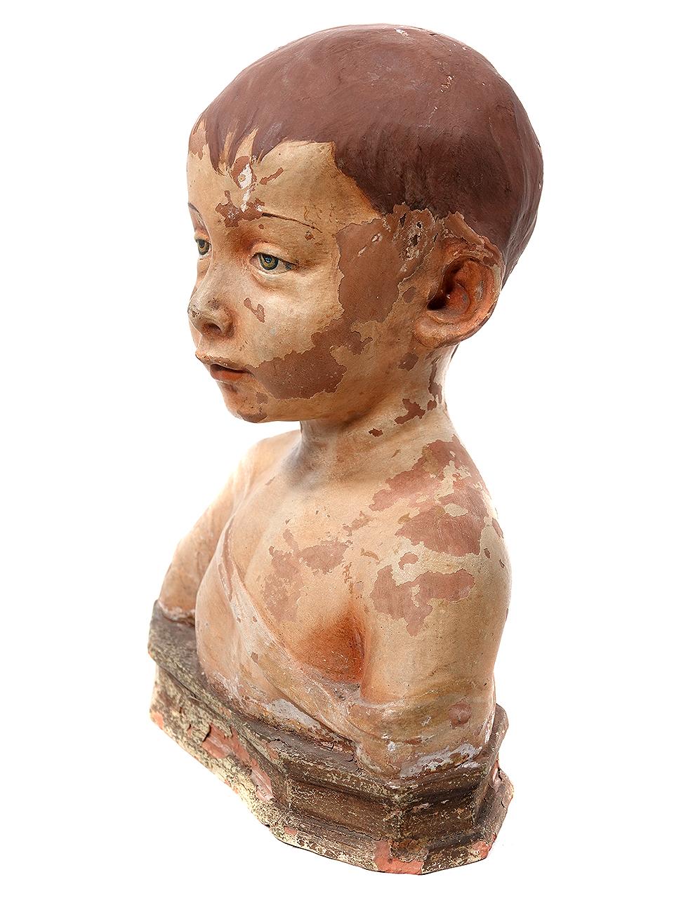 An enchanting antique bust statue of a young boy dating from the early 1900s. I am guessing on the date but it may be much earlier. The figure has been cast from terracotta and features hand-painted accents. It depicts a small child wearing a cap
