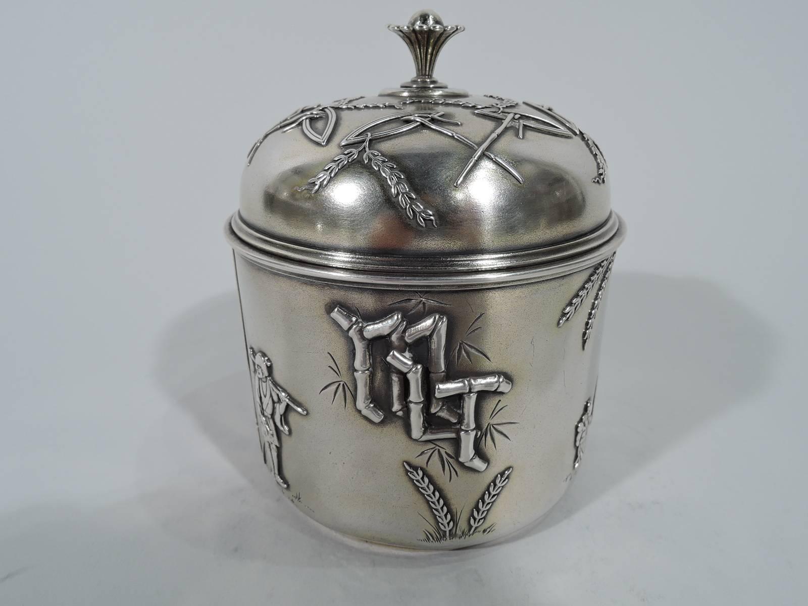 Japonesque sterling silver tea caddy. Made by Tiffany & Co. in New York. Drum form with domed cover and lotus finial. Ornament applied to butler-finished ground: Modishly exotic figures, including a parasol-carrying geisha, barelegged farmer, and