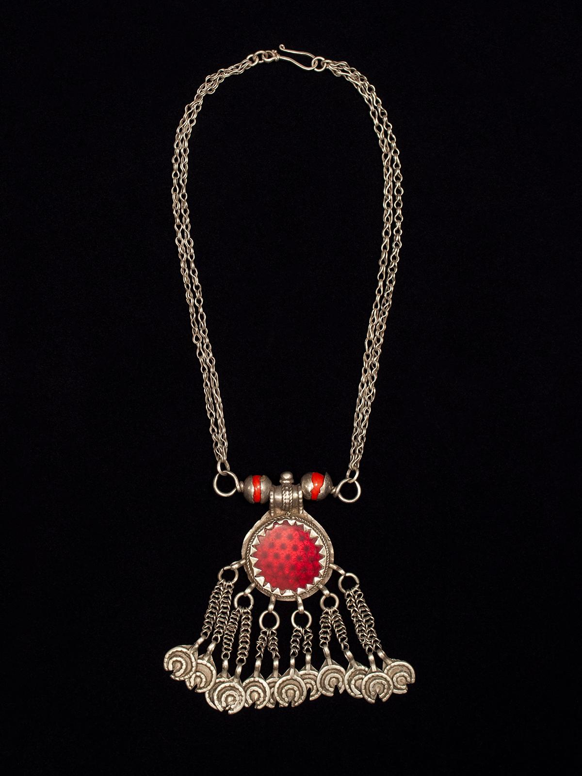 Early to mid-20th century pendant silver necklace, Yemen

This pendant necklace has an interesting center medallion of transparent red glass over a faceted sheet of silver, which gives it a reflector-like appearance. The two orange beads are wound