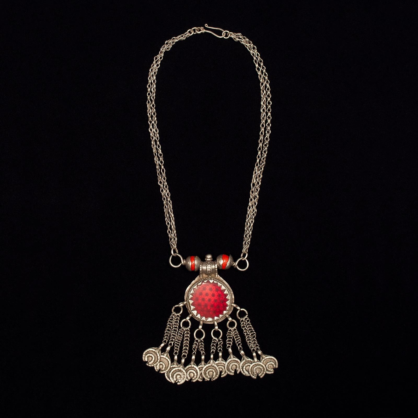 Early to mid-20th century pendant silver necklace, Yemen

This pendant necklace has an interesting center medallion of transparent red glass over a faceted sheet of silver, which gives it a reflector-like appearance. The two orange beads are wound