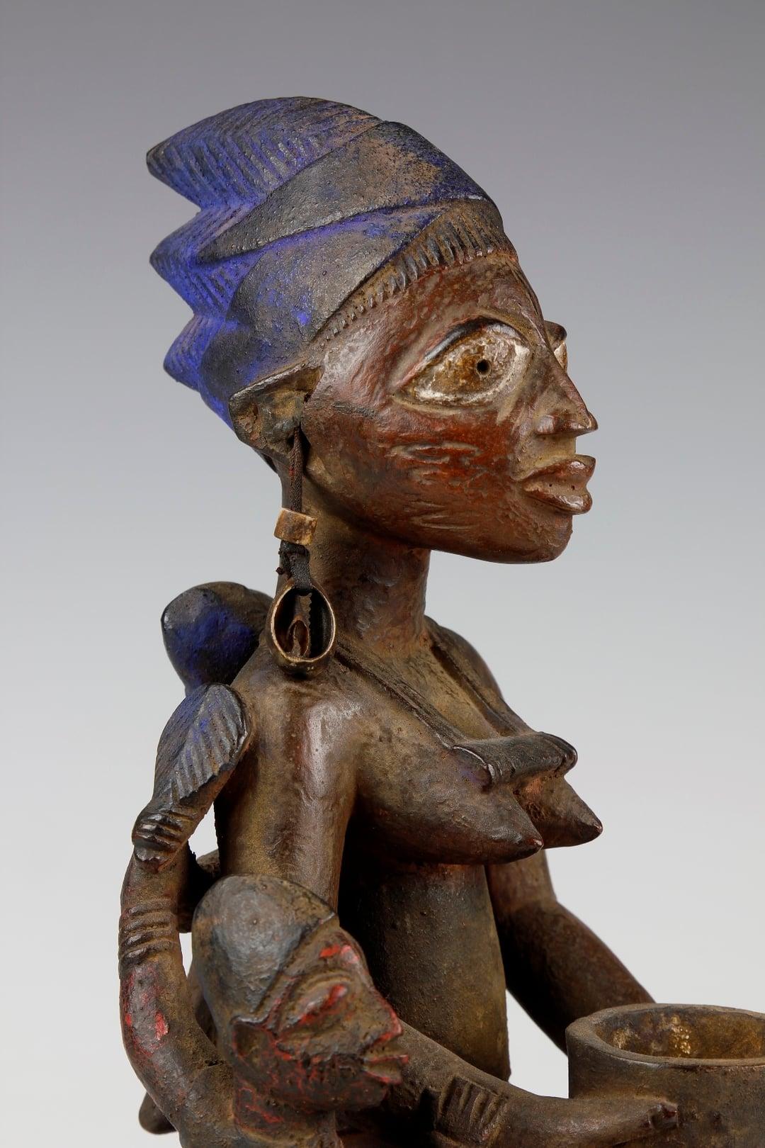 This finely carved early twentieth-century figure, from the Yoruba culture in Nigeria, depicts a mother and her two children. The mother is depicted in a kneeling position, holding a two-legged offering bowl. Crowning her head is a three-pronged