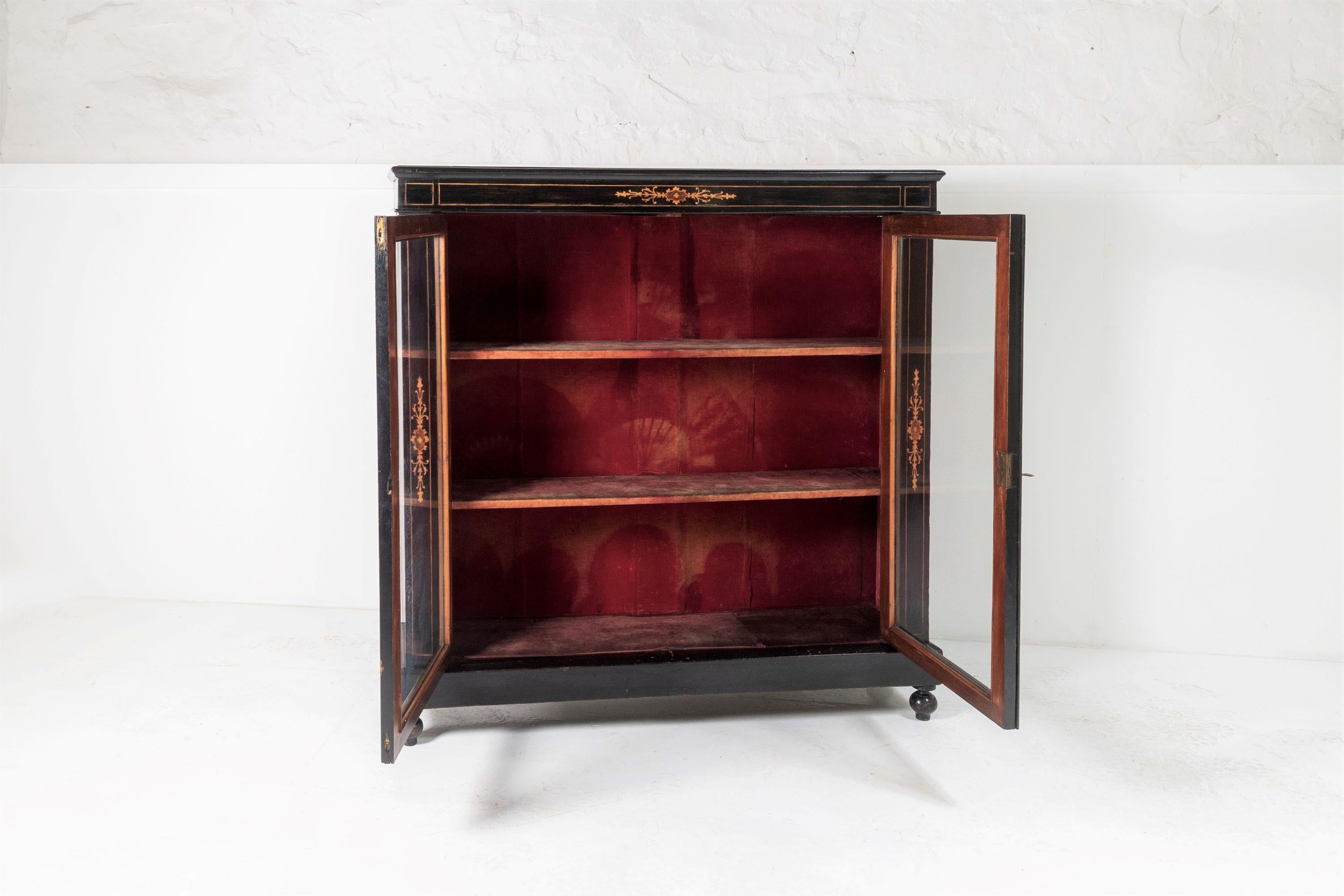 A well proportioned Victorian peer cabinet, ebonised with fruitwood inlay and standing on four turned bobbin feet.
Two opening glazed doors, retaining its original lock and key, opening to interior shelves, fully lined with original red velvet
