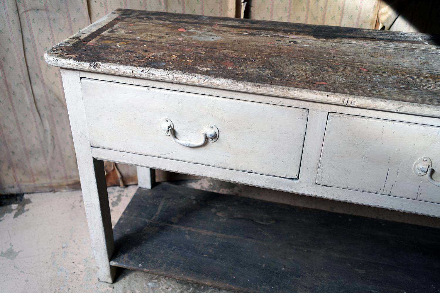 The good thick deal pine dresser base, showing a white paint distressed commensurate with age, having a characterful thick and well-worn top over three frieze drawers with drop handles, to a well-worn pot board, the whole standing on integral block