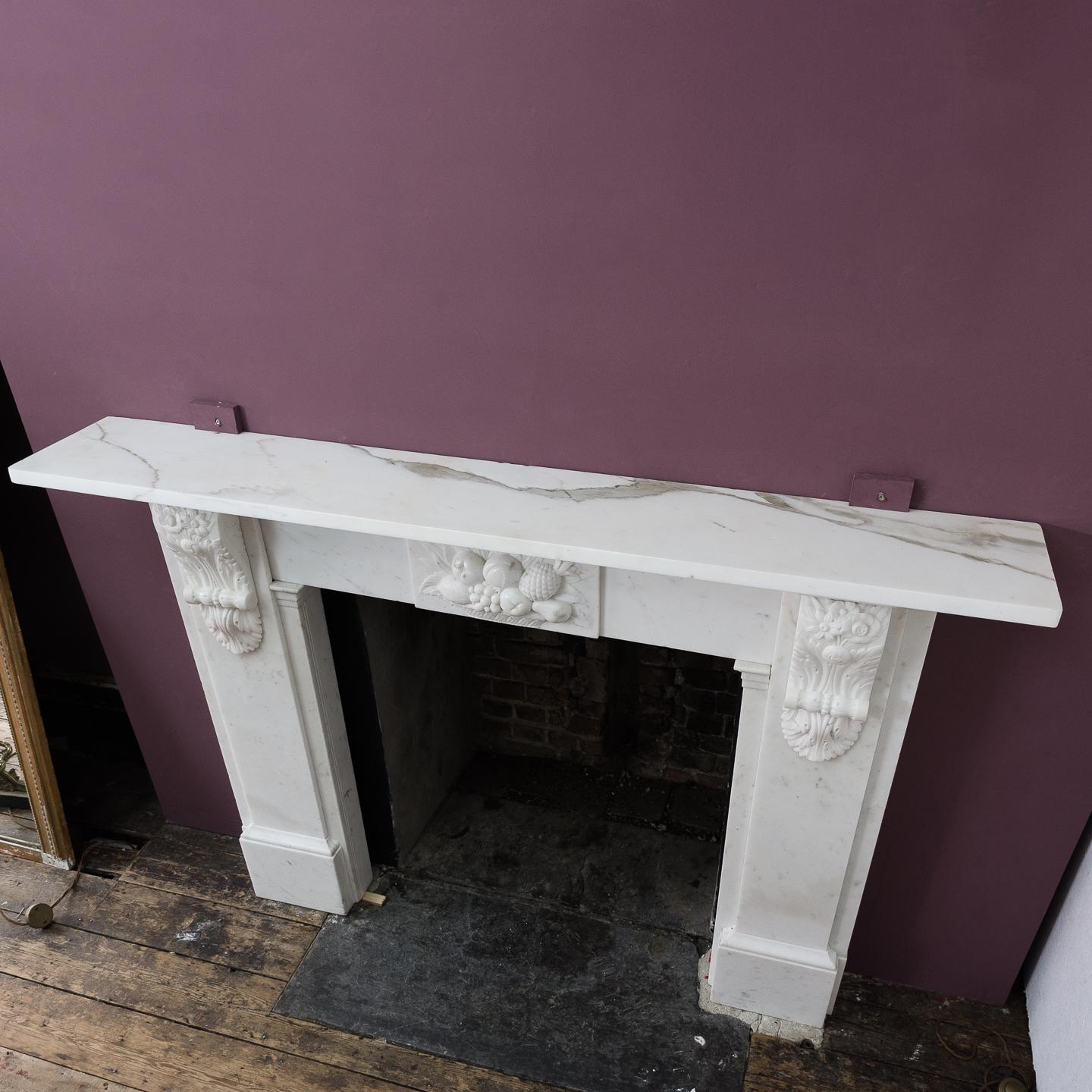 An early Victorian statuary marble chimneypiece, circa 1850-1860, the plain rectangular shelf above frieze with central tablet depicting a fruit bowl carved in relief, the plain jambs reeded by well carved foliate corbels, on foot blocks.
  