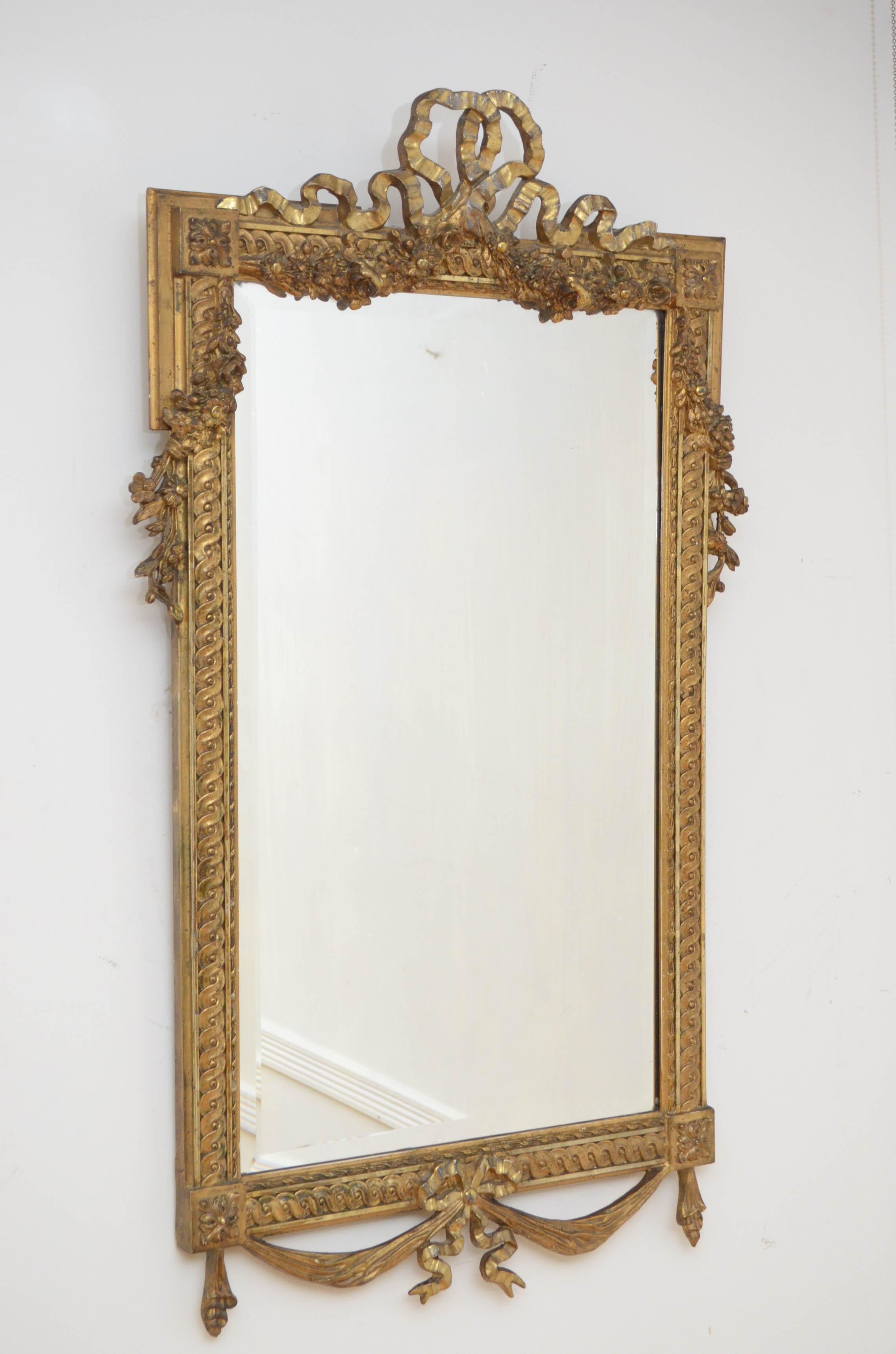 K0160 Elaborate early XXth century giltwood wall mirror, having original bevelled edge glass with some imperceptions in beautifully carved frame with swags to the base and bow crest to the top flanked by extensive floral decoration. This antique