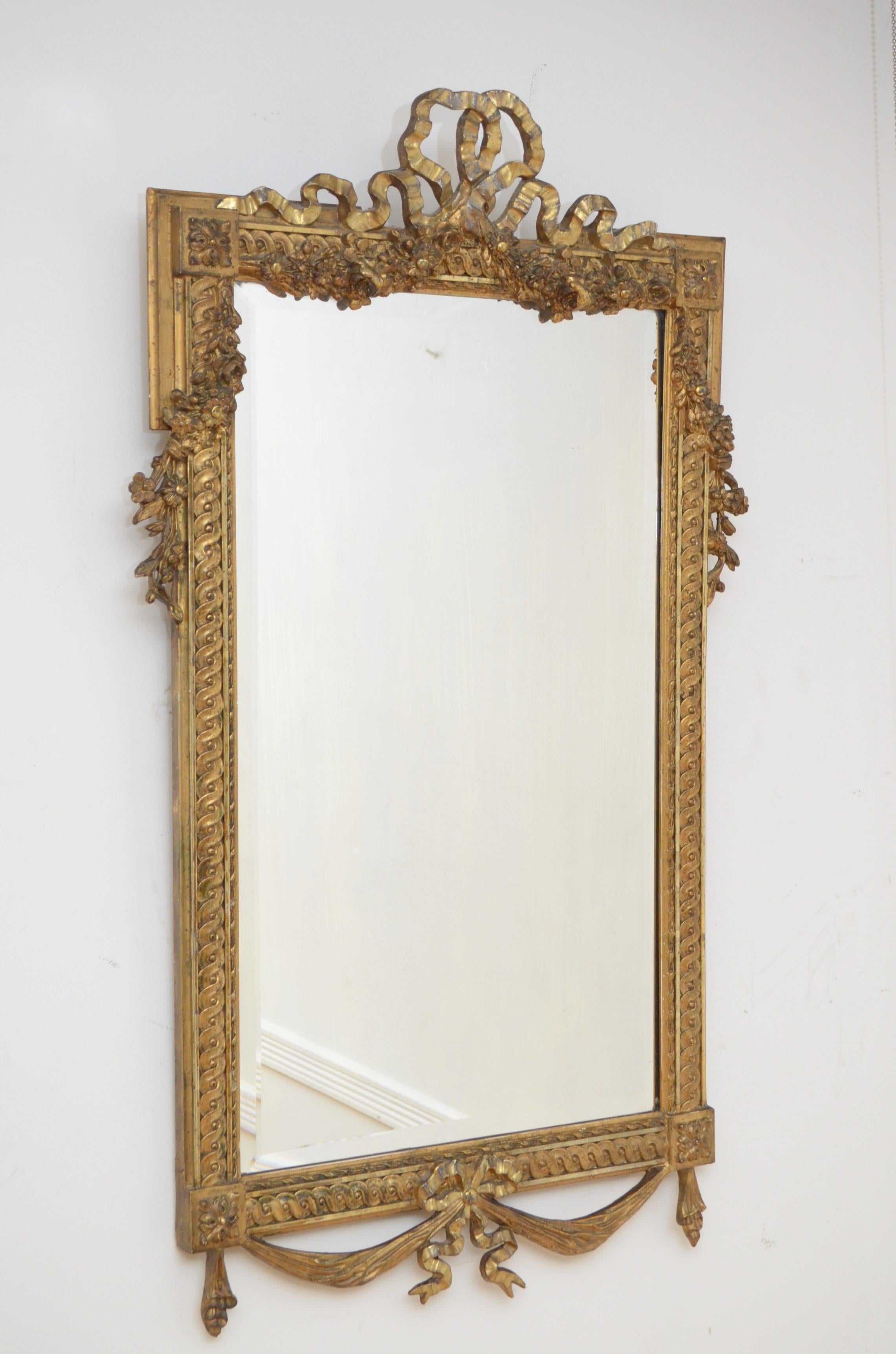 k00160 Elaborate early 20th century giltwood wall mirror, having original bevelled edge glass with some imperceptions in beautifully carved frame with swags to the base and bow crest to the top flanked by extensive floral decoration. This antique