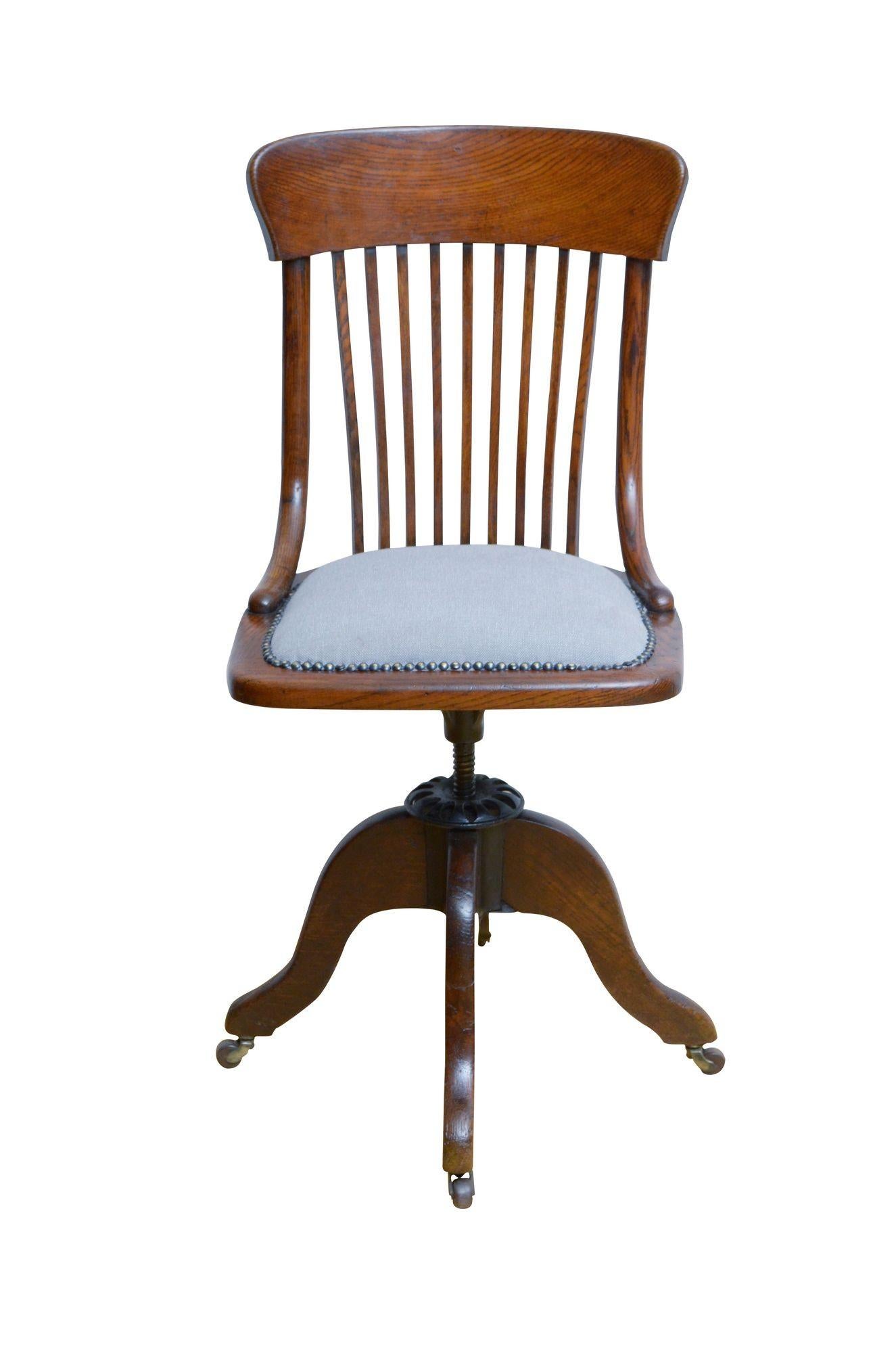 K0601 Unusual early 20th century, revolving and height adjustable chair in oak with shaped top rail, slatted back and newly upholstered closely studded seat with light grey colour top cover, standing on shaped, downswept legs terminating in brass