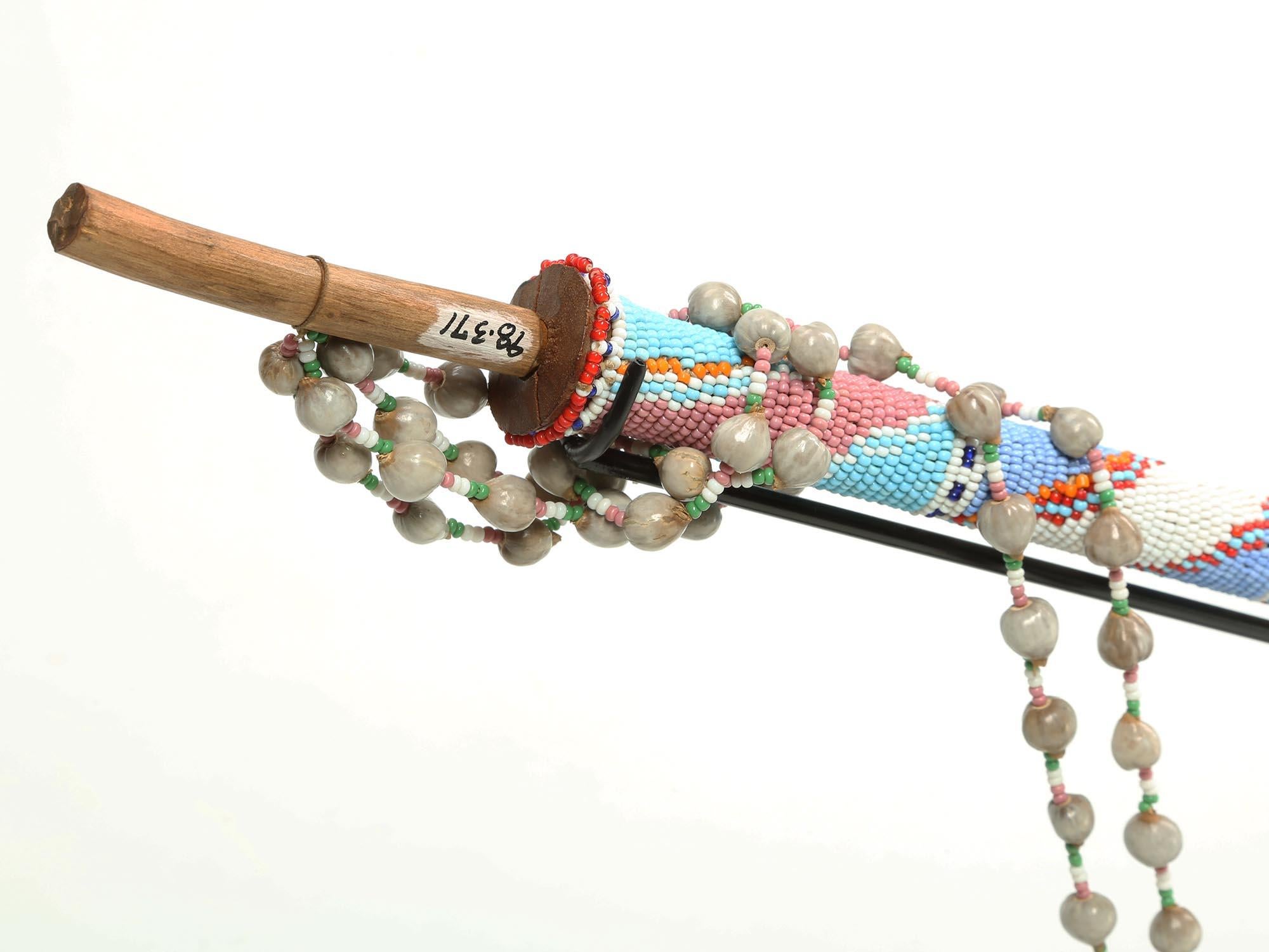 A sweetly carved wood antique Zulu smoking pipe on a custom stand. From a museum collection. Wood with glass-beaded stem and strap. South Africa. Long well carved stem under the beads with flaring mouth end, metal insert to protect wood of