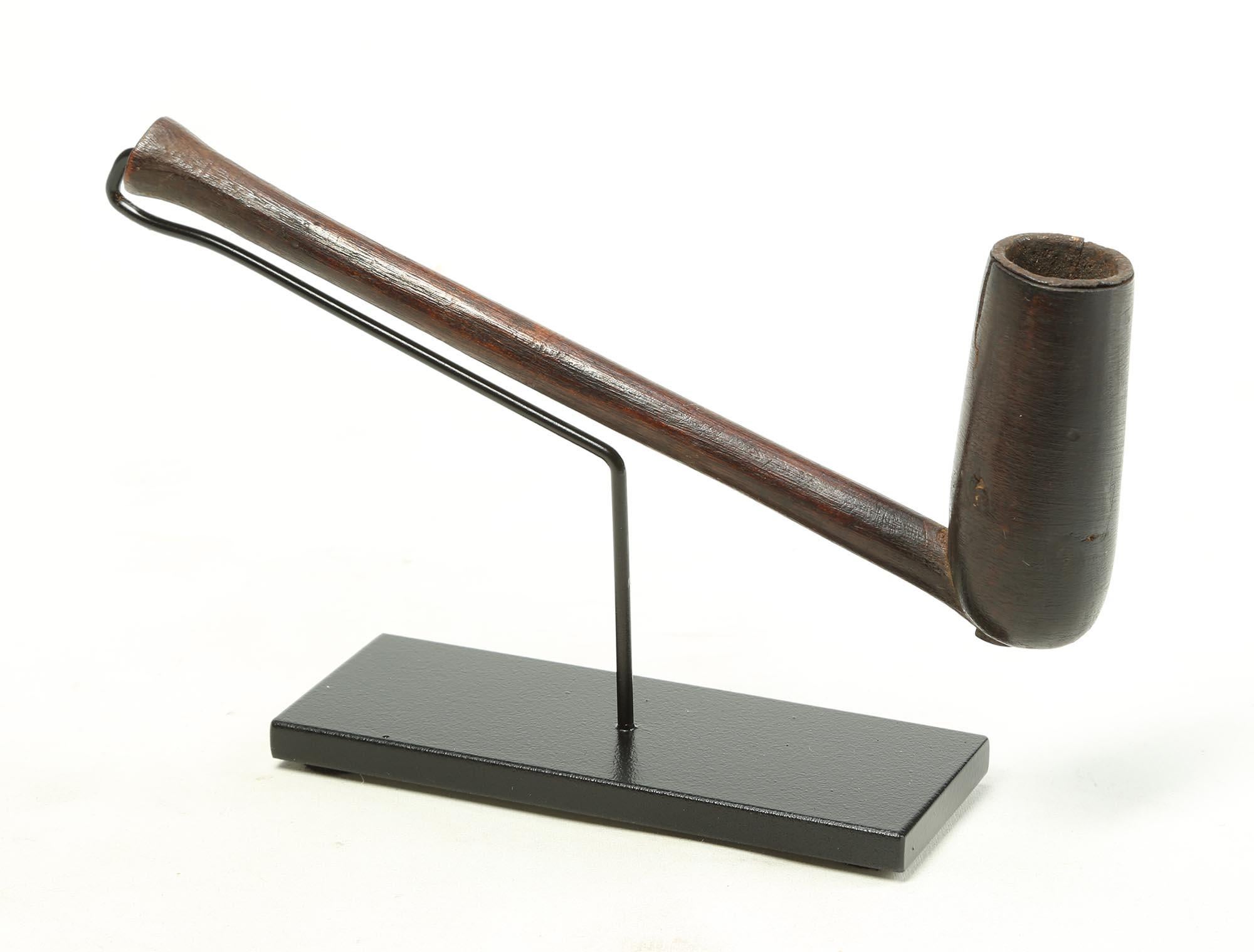 A smaller and finely detailed antique wood Zulu smoking pipe, with simple form, wood, on a custom metal stand, from South Africa. Long well carved stem with flaring mouth end, metal insert to protect wood of pipe bowl. Old crack to edge of pipe