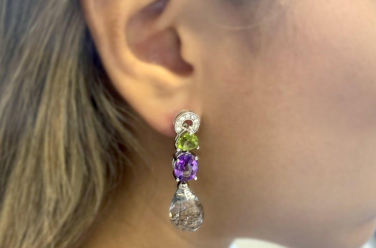 The Drop Earrings in 18kt White Gold are a captivating fusion of color and luxury. Each earring features a dynamic combination of gemstones, starting with the vibrant triangle-shaped peridot, which exudes freshness and vitality. Below, the oval
