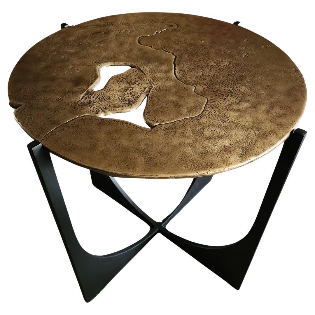 La table d'appoint Earth Prints 11 associe un plateau en laiton massif moulé à une base géométrique en acier peint en noir. La surface circulaire présente des irrégularités et des reliefs naturels laissés par le métal en fusion lors du moulage, ce