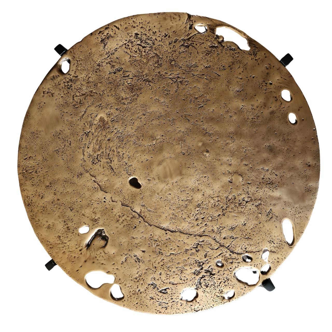 La table d'appoint Earth Prints 12 associe un plateau en laiton massif moulé à une base géométrique en acier noir. Le plateau rond conserve les marques brutes du métal en fusion - les cavités, les textures et les reliefs irréguliers apparaissent