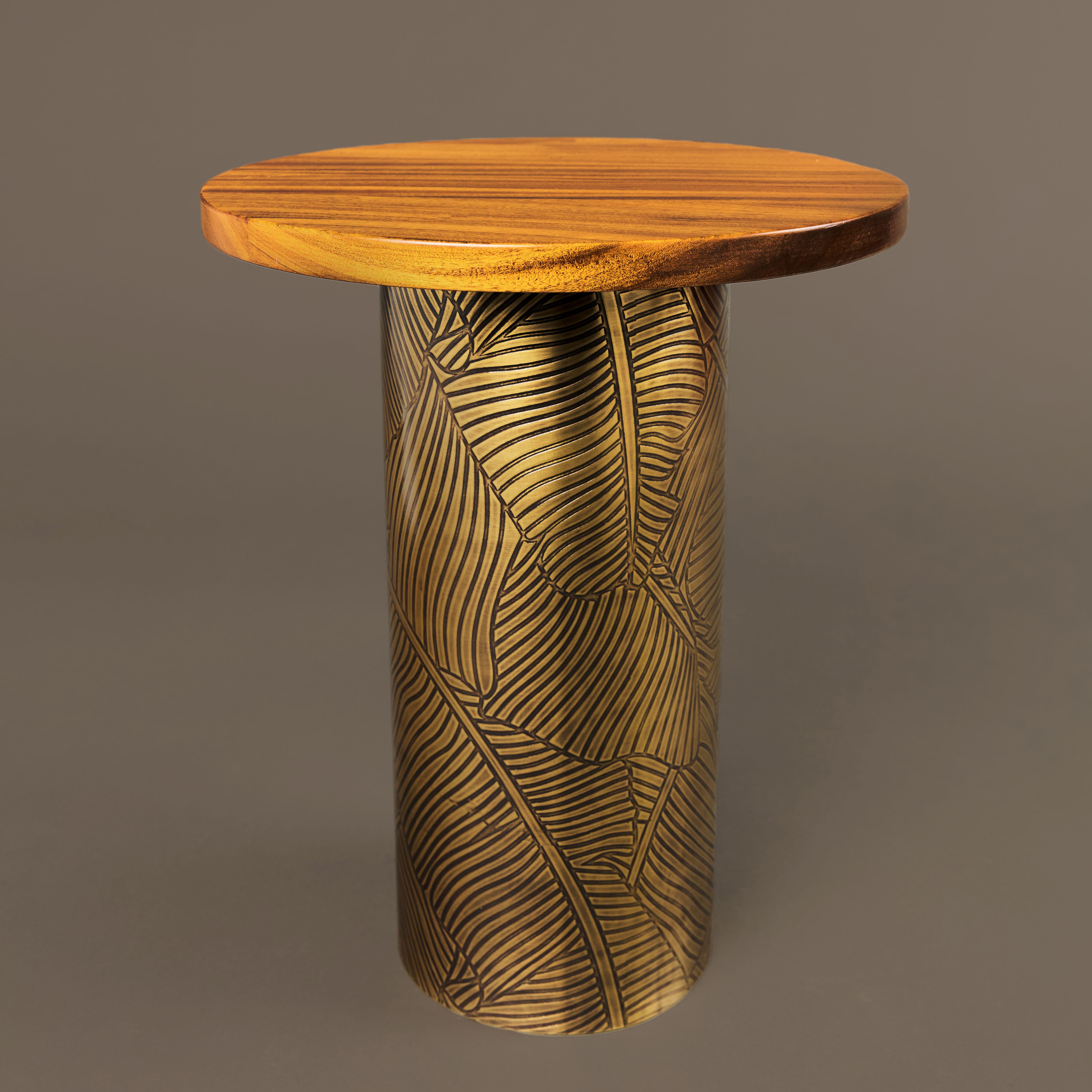 La table d'appoint Earth Prints 6B présente un plateau rond en bois de dibétou massif posé sur une base cylindrique en laiton gravé, finie avec une patine chaude d'or vieilli. Le motif gravé, inspiré de formes botaniques stylisées, se déploie en