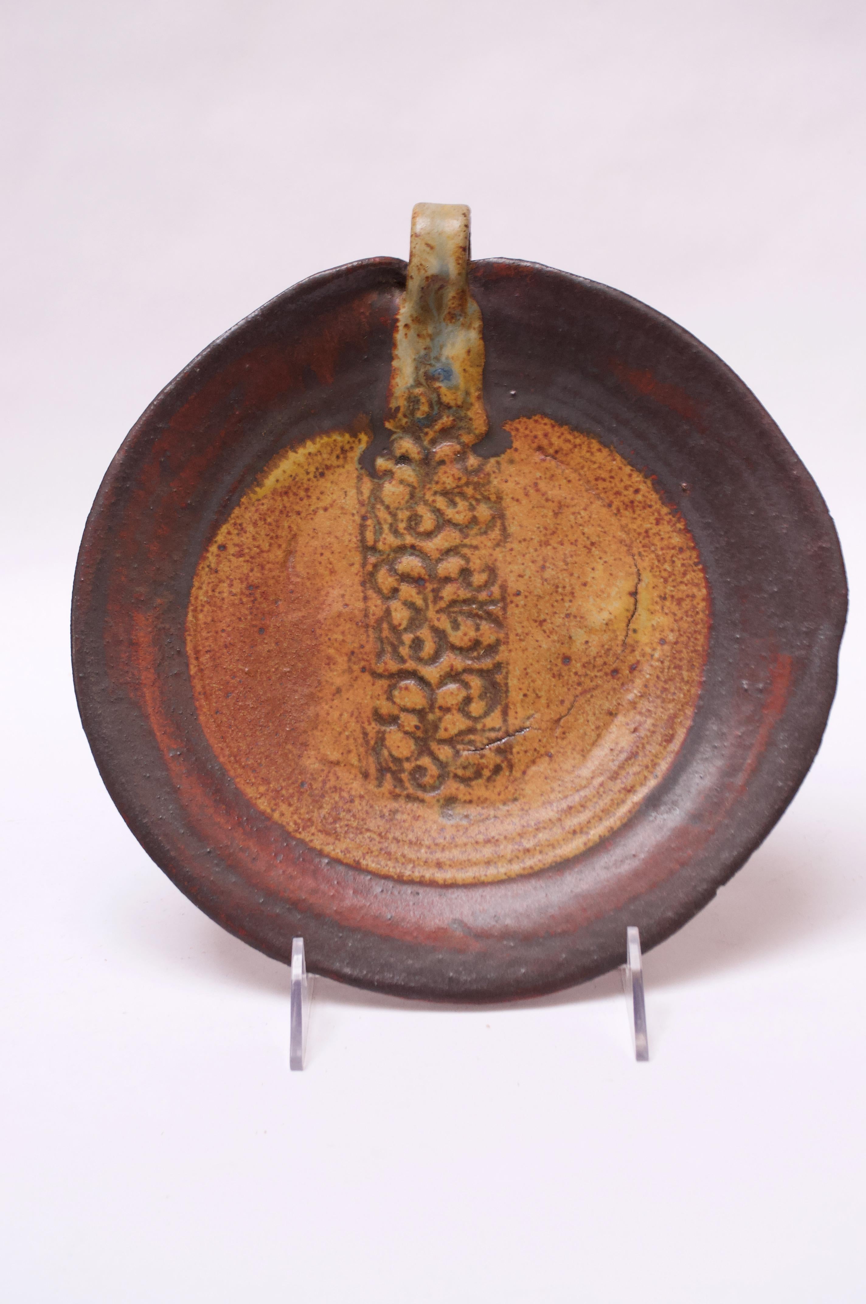 Striking stoneware charger / decorative plate in attractive earth tones of ochre and brown with swaths of red and a strip of brown leather attached to the handle. Central pattern featuring sgraffito technique, adds a nice textural element, while the