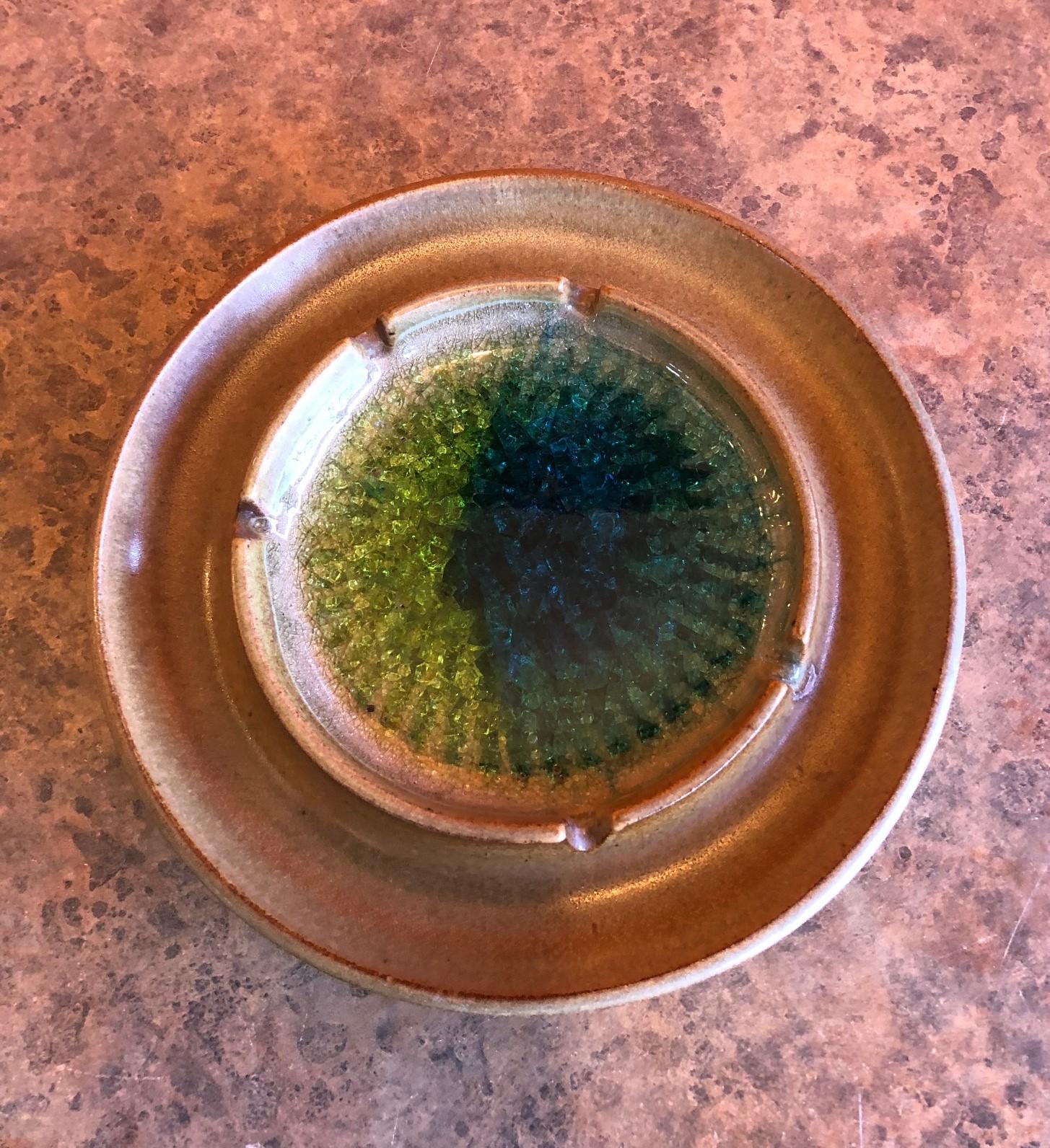 A wonderful earthenware ashtray with green, yellow and blue Blenko crackled glass in the center signed by Robert Maxwell, circa 1960s. The piece measures 8