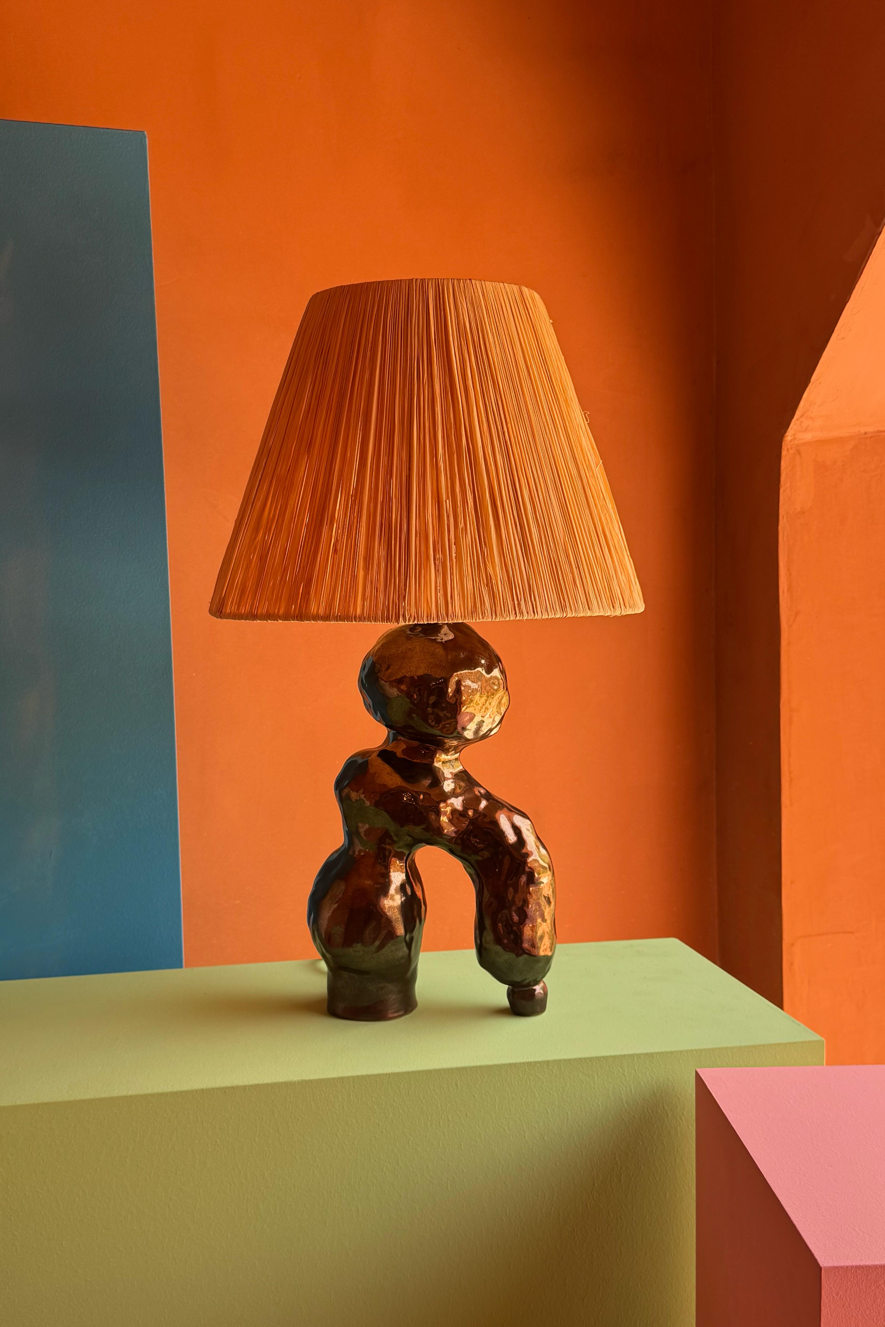This hand built, ceramic lamp is made from earthenware and glazed in the artist's own 	glaze, giving the body of the lamp a metallic patina. The raffia lampshade has been handmade by the artist. 

All elements are the creation of the artist in his