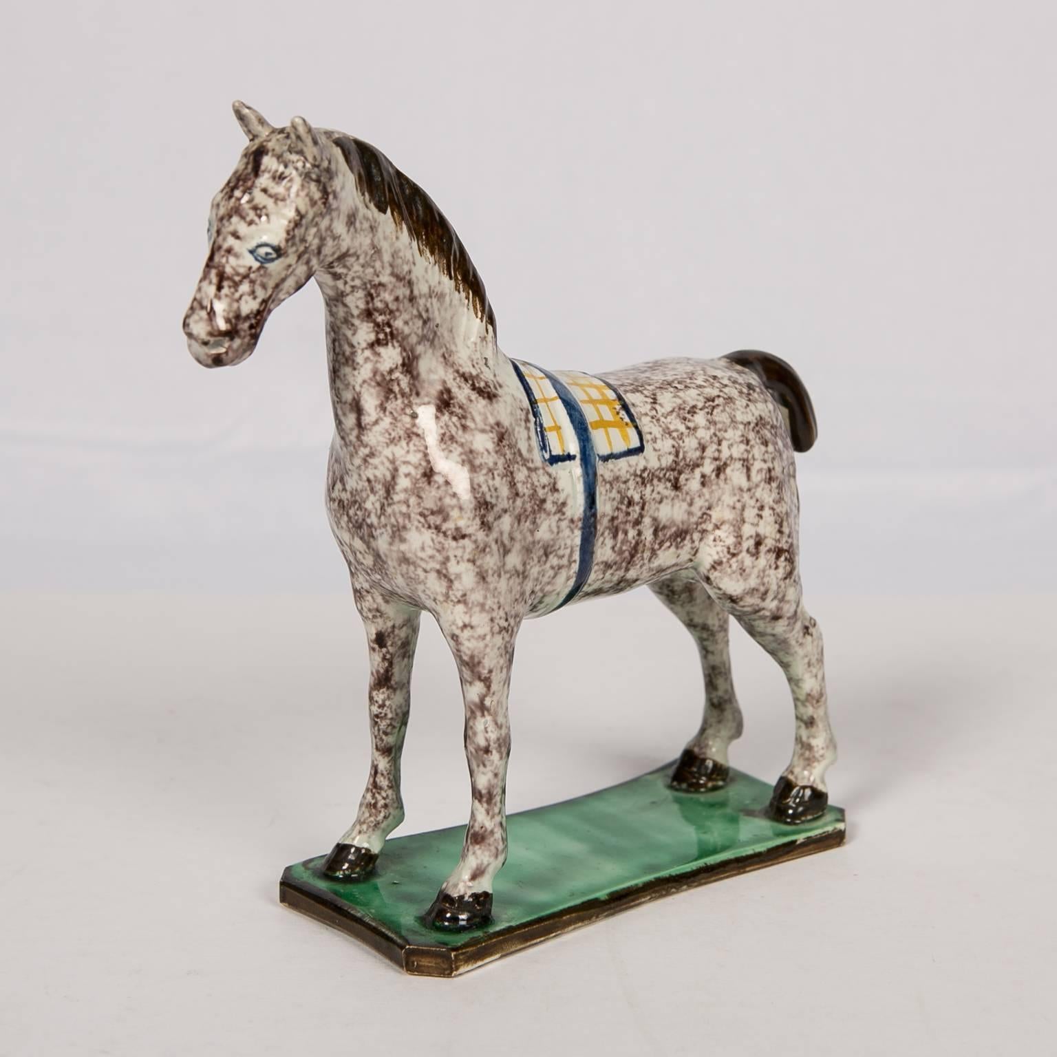 A naive figure of a pearlware horse standing on an oblong grassy green base. The figure is well modeled and simply decorated with a sponged decoration replicating the coat of a dappled gray. The artist painted a saddle blanket of white and yellow