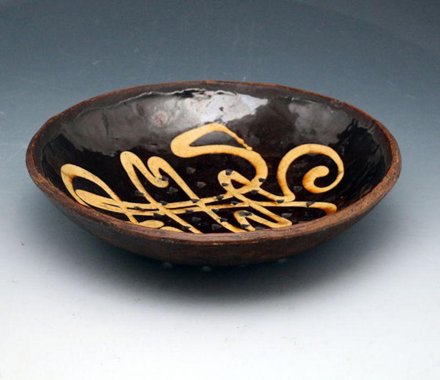 A fine earthenware circular shaped dish or colander with a dark chocolate ground decorated with an elaborate calligraphic honey colored trailing slip. The dish has been made with a center grouping of 