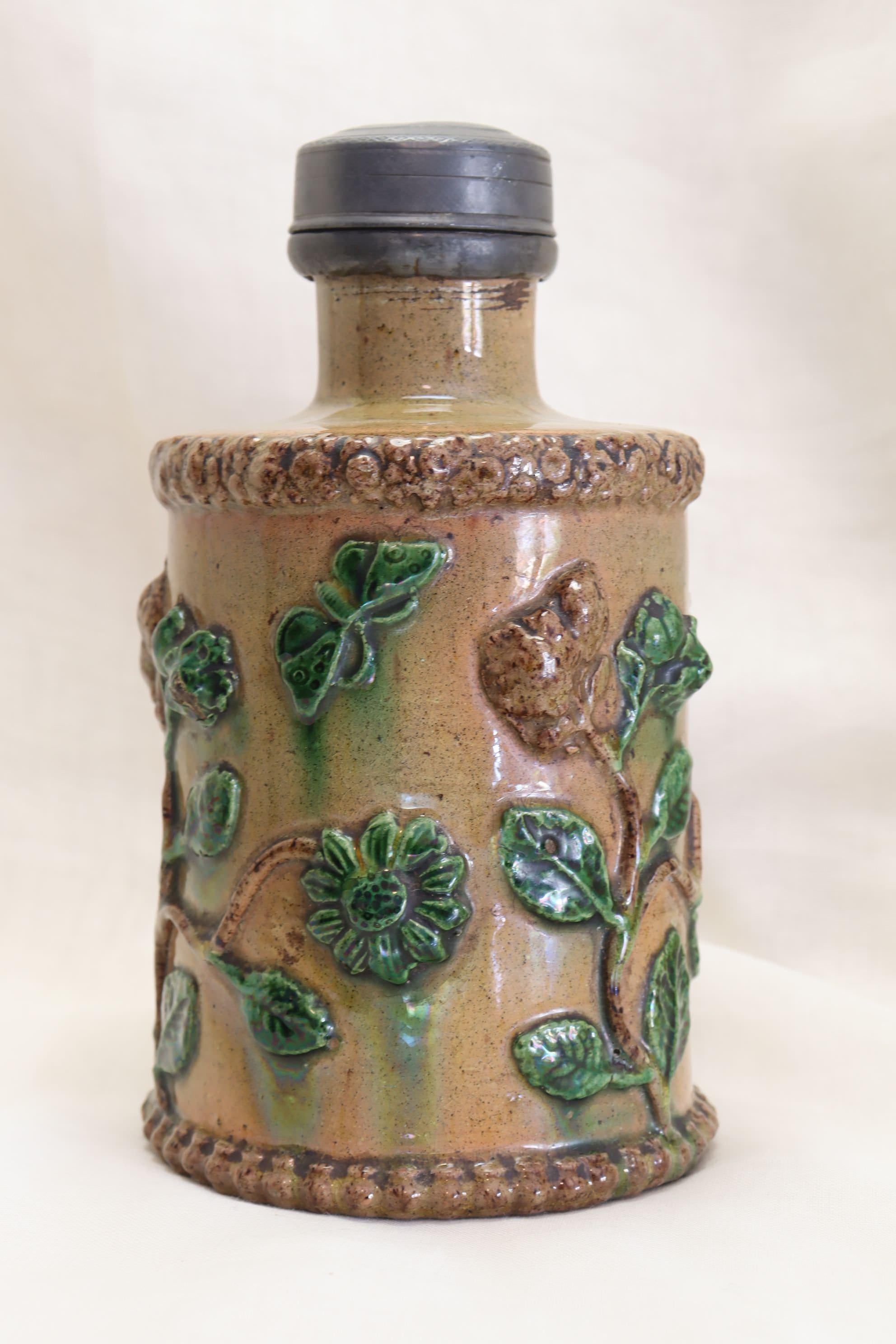 This pottery tea canister is decorated with very detailed, and quite pronounced, applied sprigs of fruit, flowers, leaves and butterflies, and there is a slight iridescence to the glaze. It is capped with a screw on pewter lid which has a very