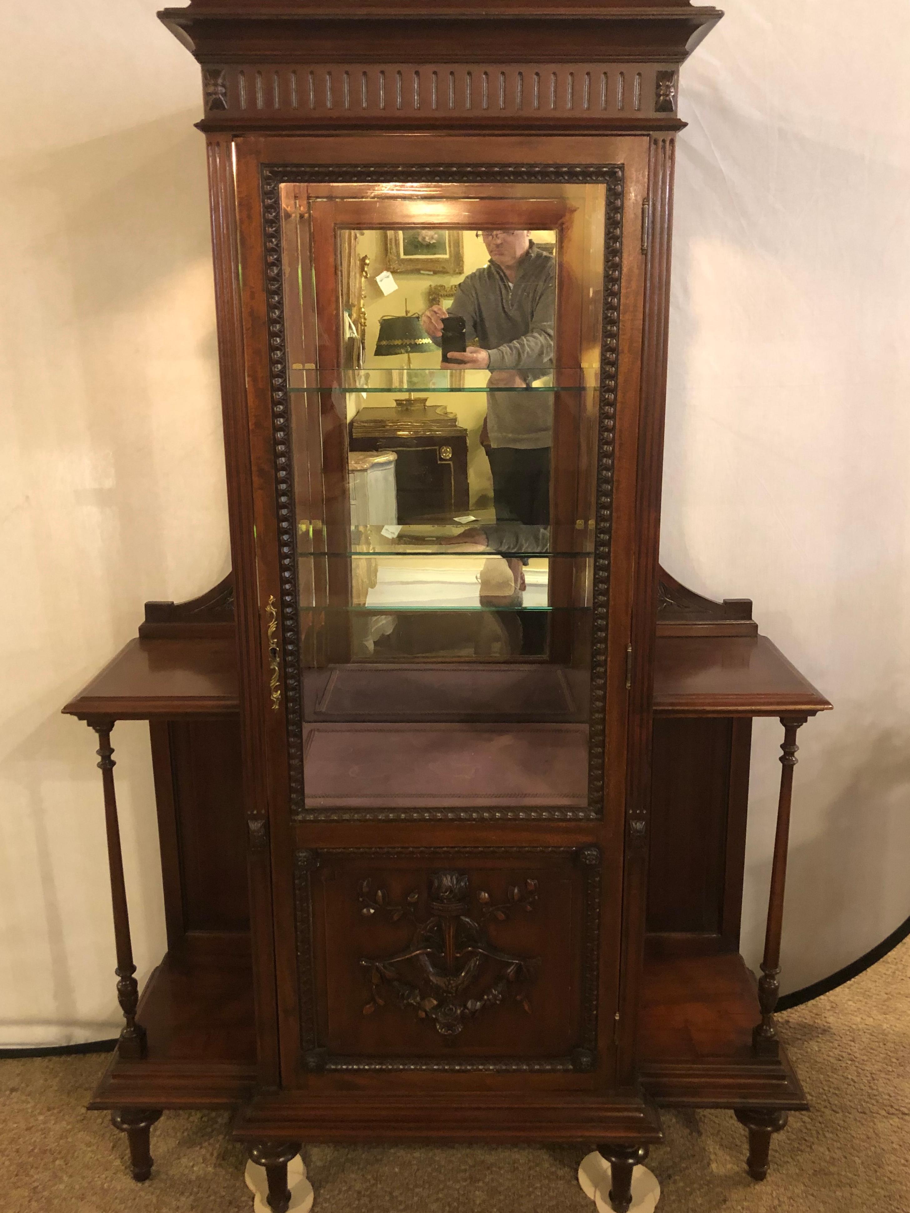 An East Lake Victorian 19th century Vitrine or curio cabinet having a mirror back with glass shelves. This vintage piece is in very fine condition and comes with a lighted interior. The centre door with a wonderfully detailed carved solid wood torch