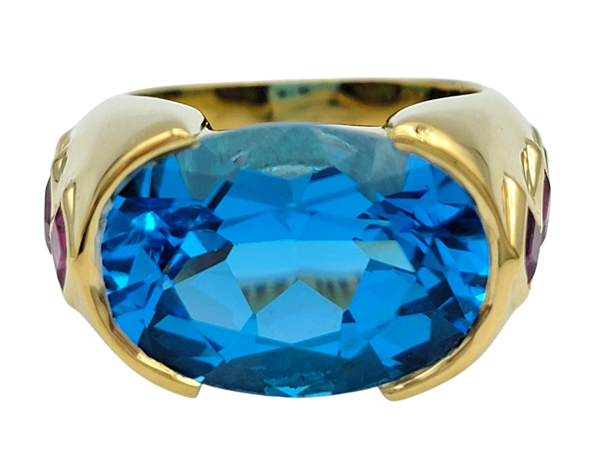 Ring Size: 8

This eye-catching ring features a bold oval-cut Swiss blue topaz set horizontally across the finger. The vivid blue gemstone takes center stage, framed by a checkerboard of channel-set princess-cut rubies in rich red tones along the