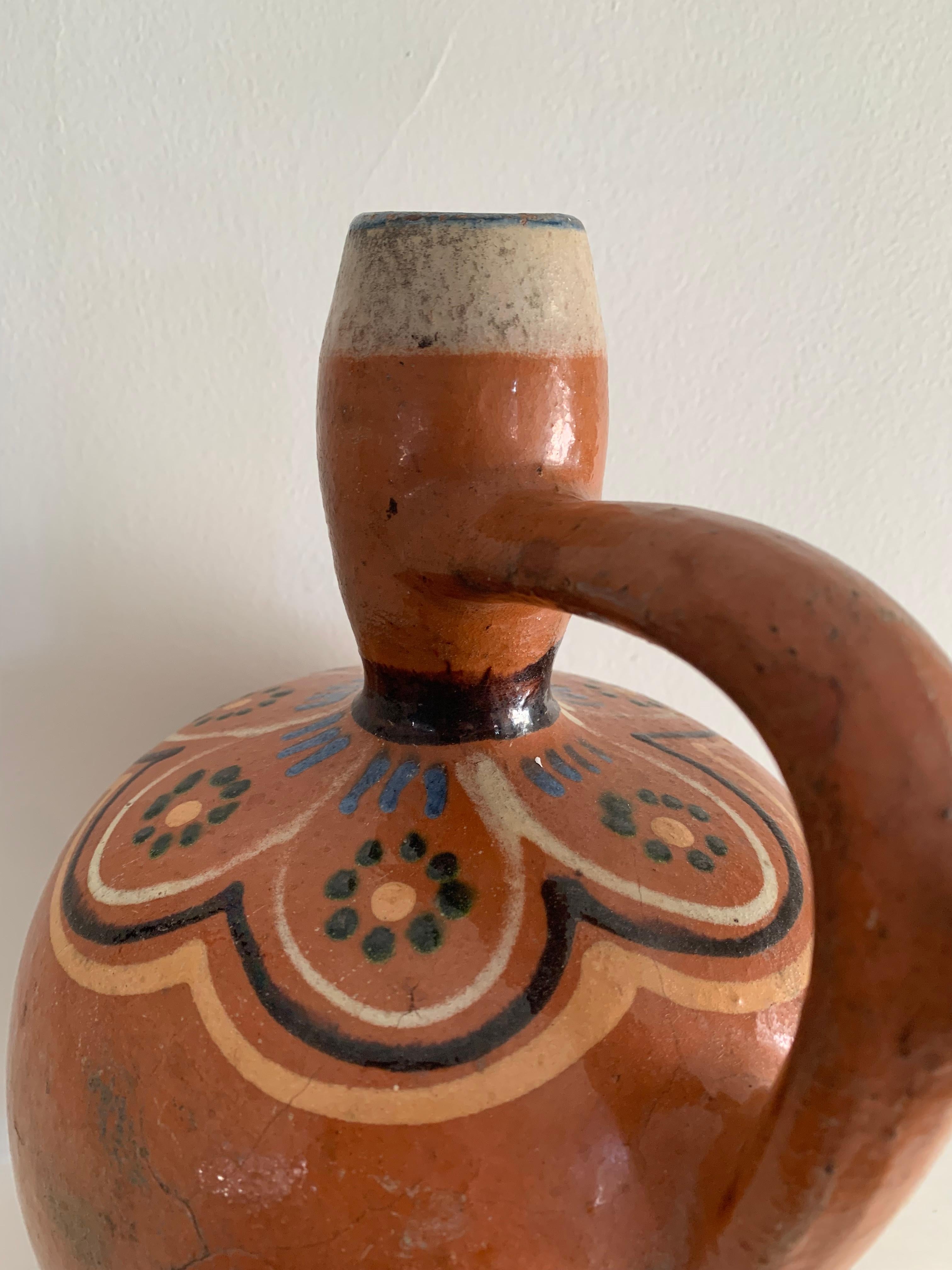 Rustic hand-thrown earthenware jug with bulbous body, tall neck, and expressive pulled handle. Decorated in ochre, black, green, and cobalt slip beneath a transparent glaze, forming a stylized scalloped floral motif around the shoulder. The red clay