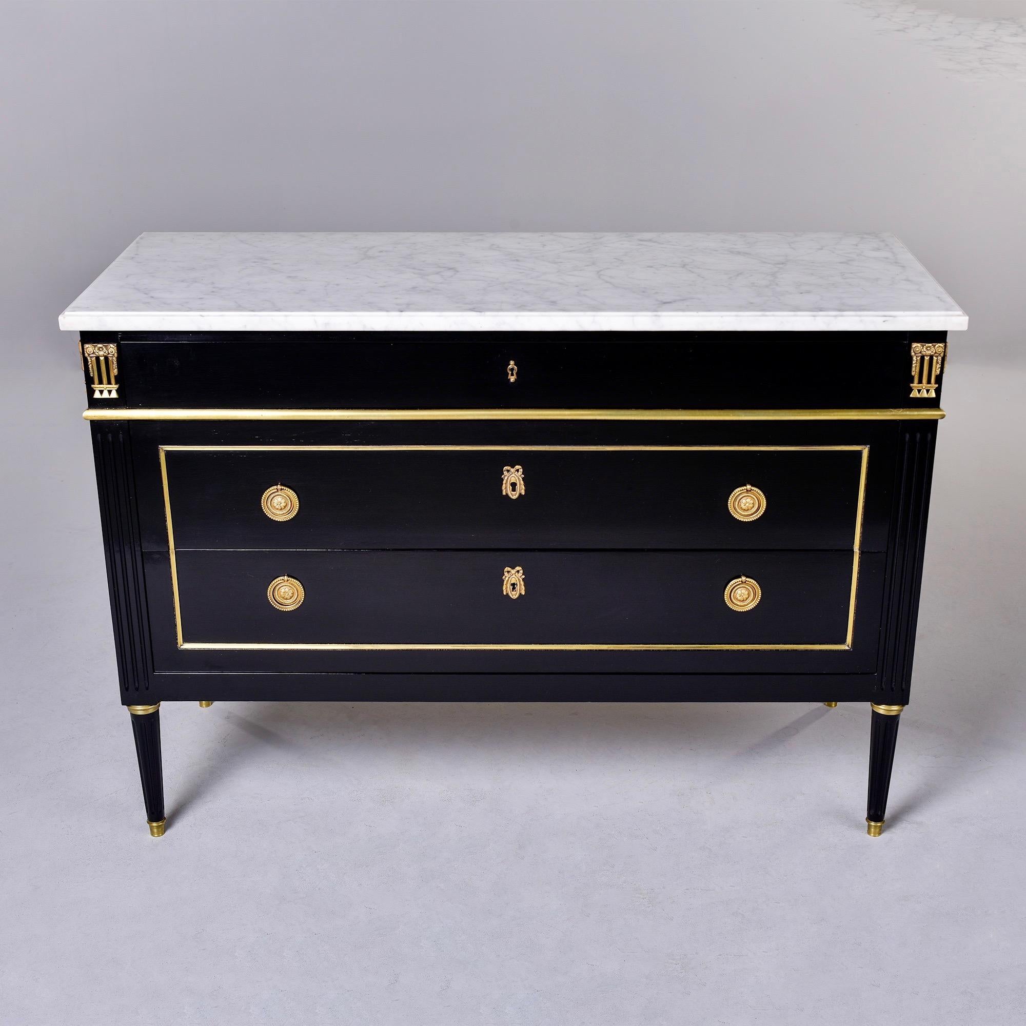 Circa 1900 Louis XVI style commode of ebonised mahogany with brass mounts and white marble top. Commode has a narrow locking top drawer with decorative brass mounts and two larger drawers below that lock with a skeleton key. Brass trim on drawer