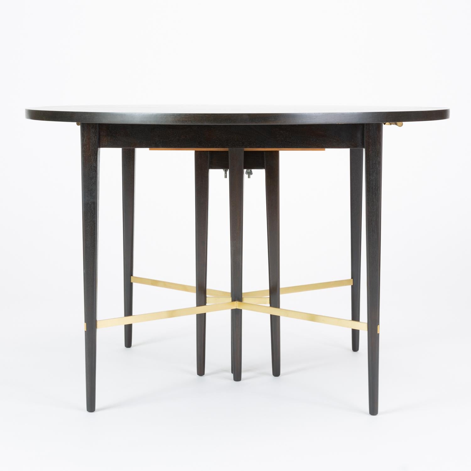 A round dining table from the 1954 Calvin Group, designed by Paul McCobb for Calvin Furniture. This example has been ebonized, emphasizing its slender lines and the dynamic angles of the legs and polished brass stretchers. The table has six 12.75”