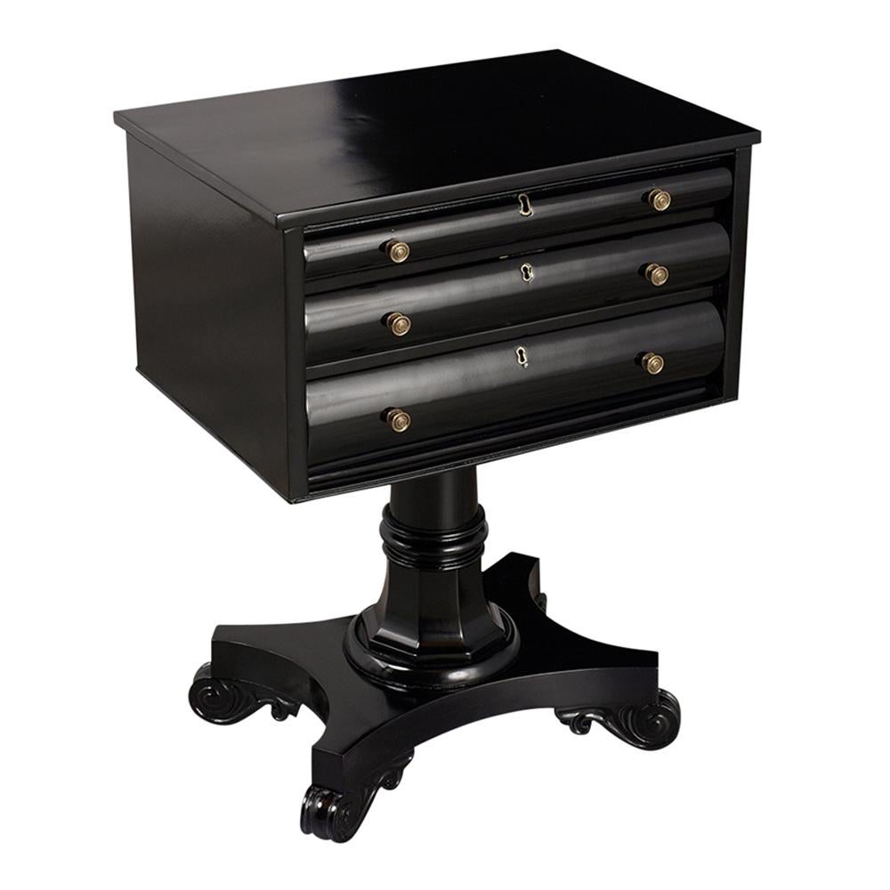 This 1840s Regency Empire Style Mahogany  Side Table has been fully restored and has a new ebonized lacquered finish. It features three drawers, each with a lock and brass knobs. and rests on a raised pedestal legs with hand-carved scroll design.