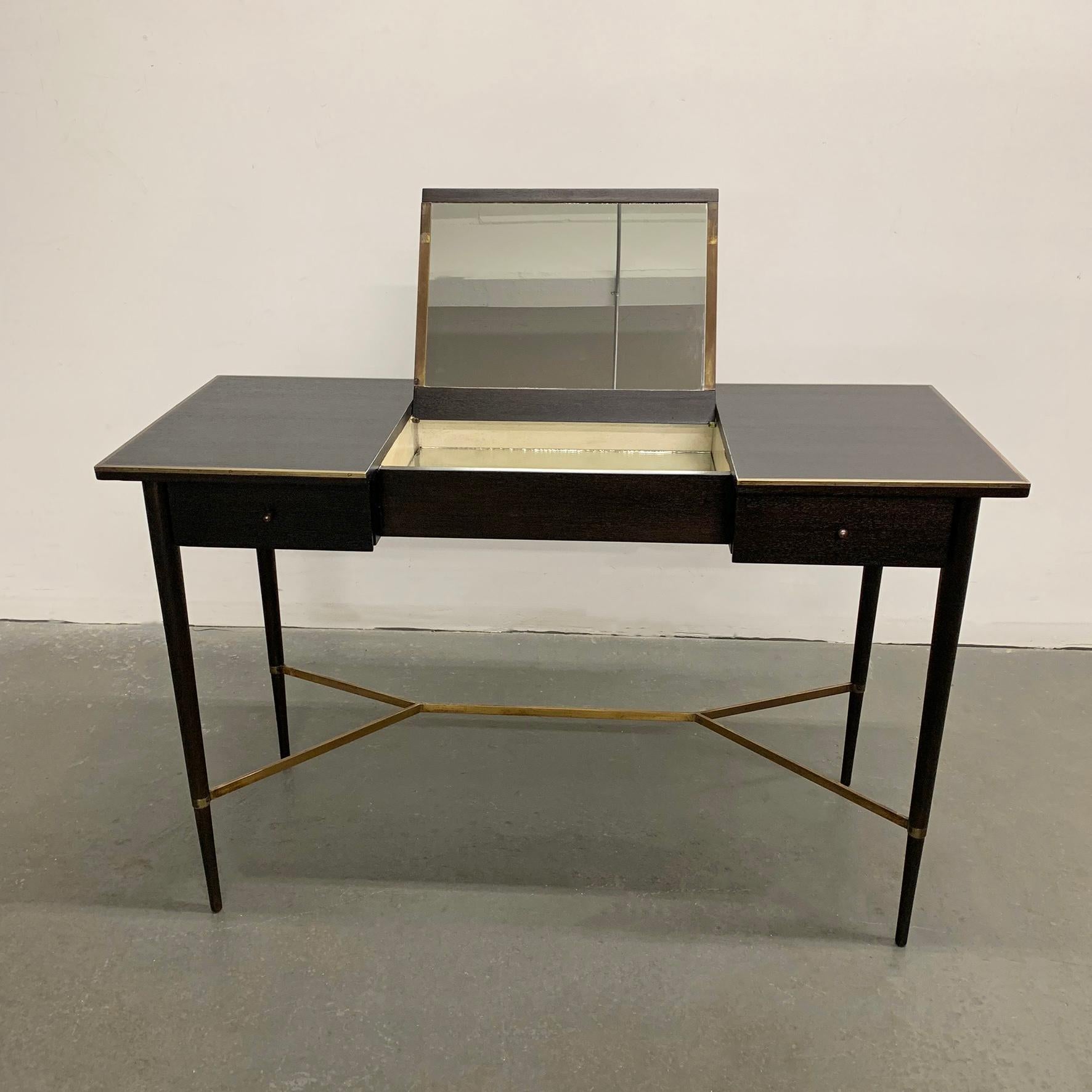 Mid-Century Modern, ebonized mahogany vanity table by Paul McCobb for Calvin, Irwin collection features beautifully tapered legs with brass stretcher, brass trim on top and 2 drawers with signature, hourglass, brass pulls. The vanity opens on top to