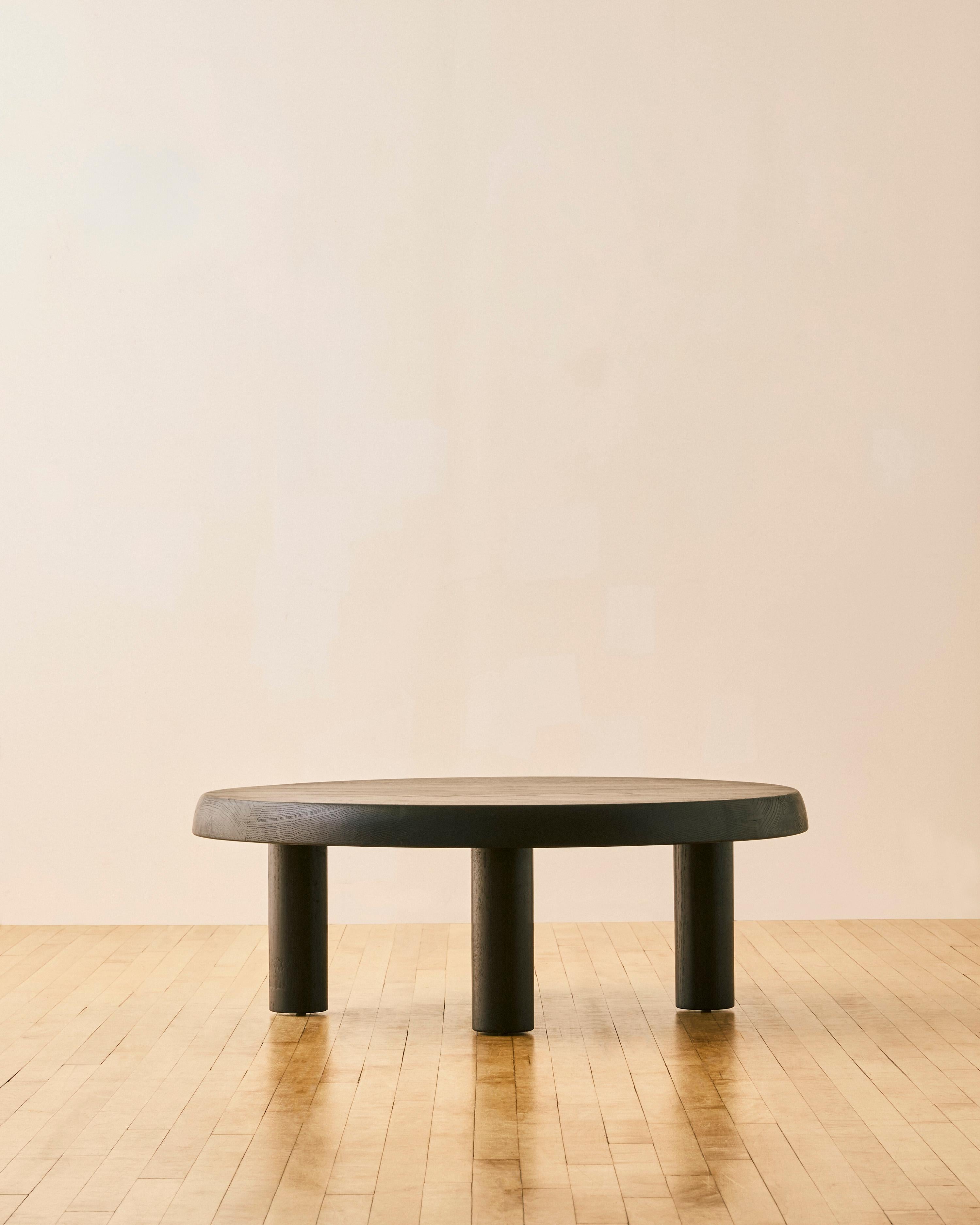 Mesa de centro de roble ebonizado a la manera de Charlotte Perriand, con tablero redondo de losa de madera maciza y cuatro patas cilíndricas con juntas de mariposa en la parte inferior. 