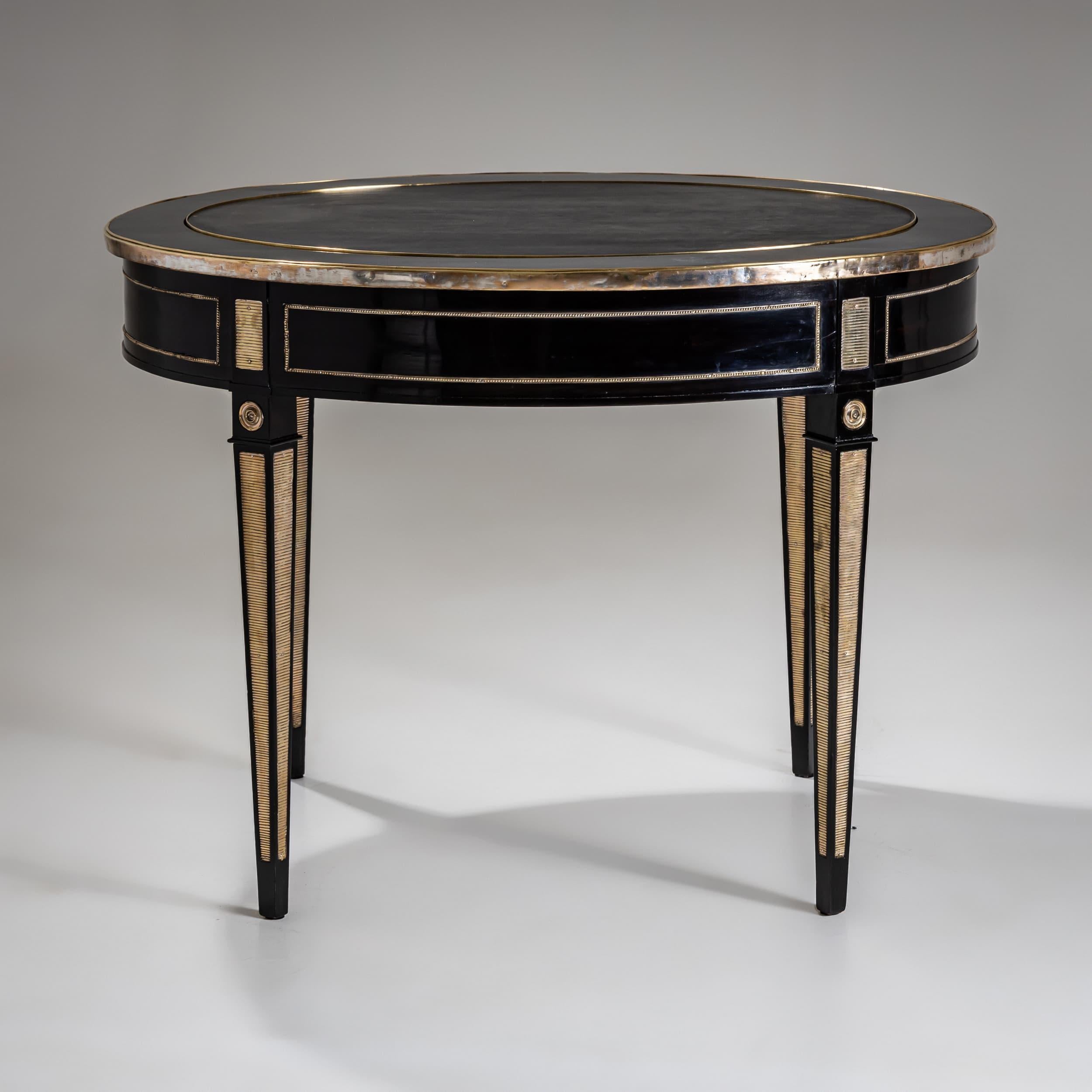 Elegant 19th-century desk with an oval tabletop, fully ebonized and adorned with finely crafted brass appliqués. The perimeter is framed by a continuous brass border. At the center, a removable dark leather-inset oval reveals a shallow hidden