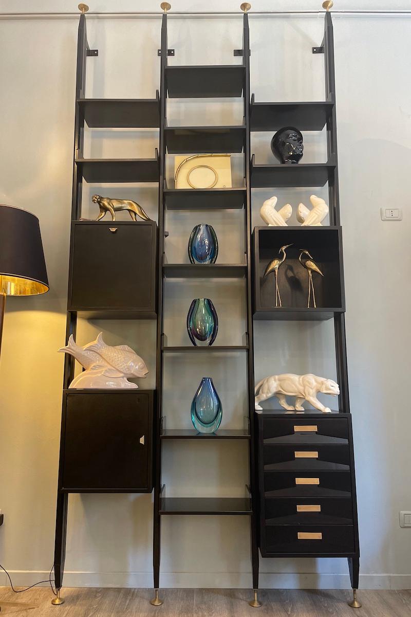 1960s bookcase in the style of Franco Albini, made of ebonized wood, featuring adjustable shelves, niches, drawers, and repositionable cabinet doors.
The structure is fully demountable and includes brass feet at both the top and bottom, typical of