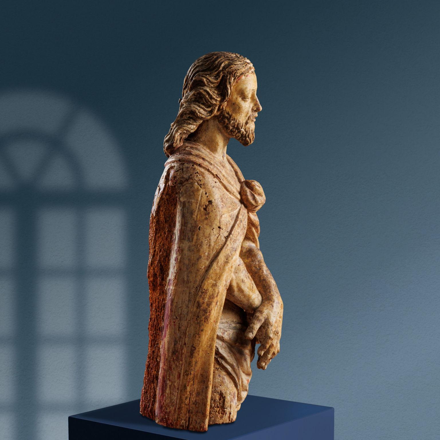 Wooden sculpture depicting Ecce Homo. Christ, whose figure is cut off at thigh level, is standing, arms crossed at waist level. Long ringlets frame a face with a pointed beard, a half-open mouth, and a low stare in an attitude of resignation. He