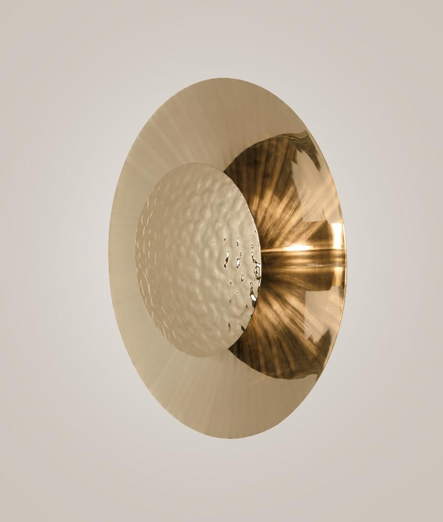 Este aplique de pared dorado presenta dos placas redondeadas de latón ligeramente separadas, que permiten que brillen pequeñas medias lunas de luz a través del hueco. La placa frontal está martillada a mano mediante técnicas artesanales modernas,