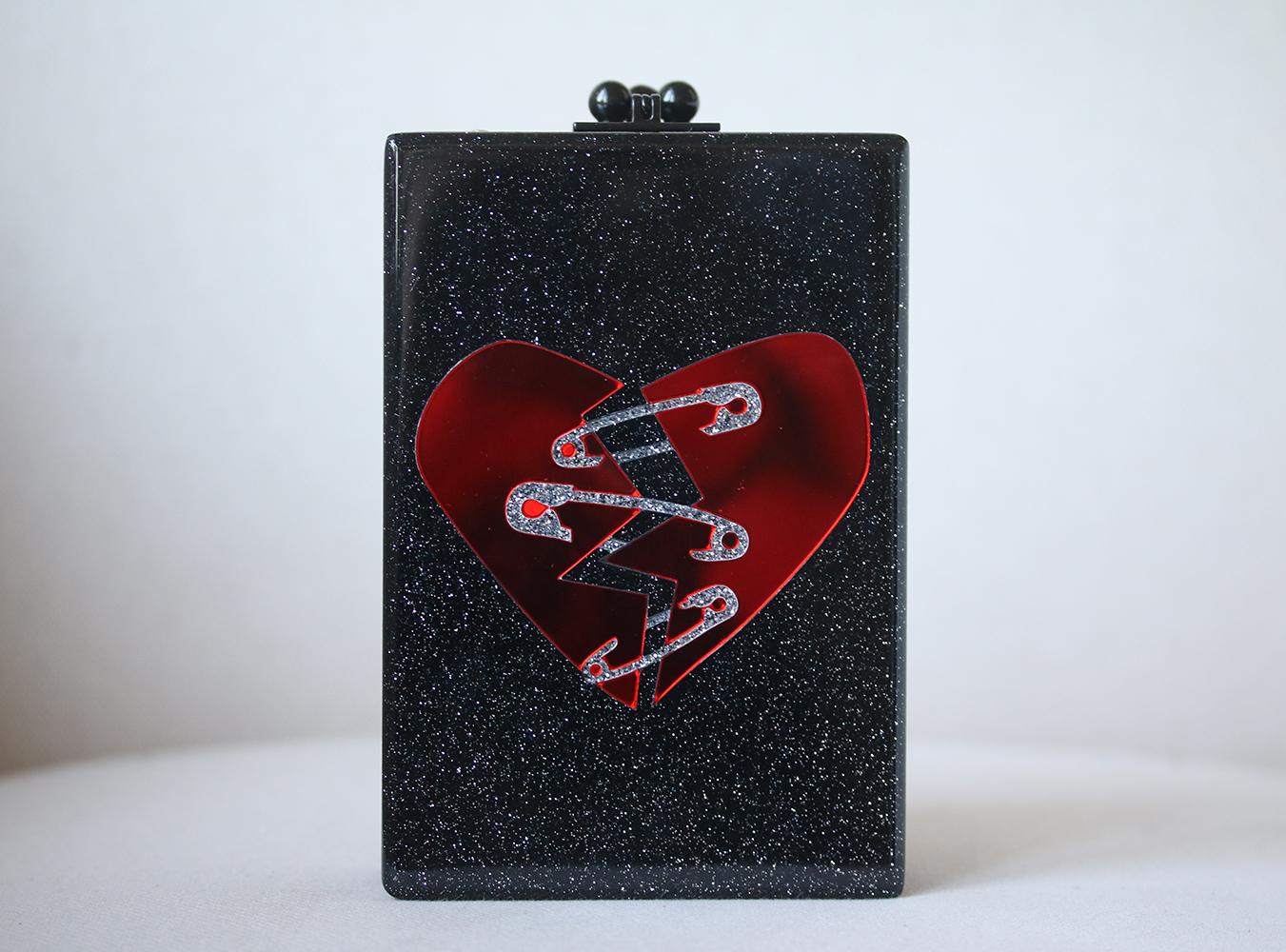 This Edie Parker Carol clutch is rendered in hand poured acrylic and features a red broken heart design and a drop in chain shoulder strap. Interior mirror with etched logo. Hand poured acrylic. Colour: Glitter black. Kiss lock closure.

Dimensions: