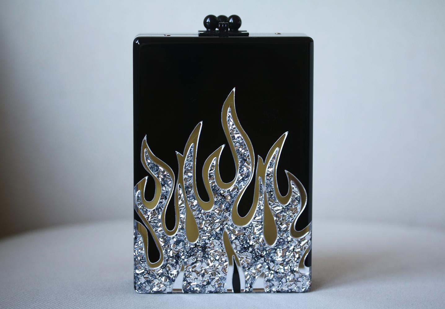 This Edie Parker Carol clutch is rendered in hand poured acrylic and features a silver flame design and a drop in chain shoulder strap. Interior mirror with etched logo. Hand poured acrylic. Colour: Black. Kiss lock closure.

Dimensions: H 16.2 cm x