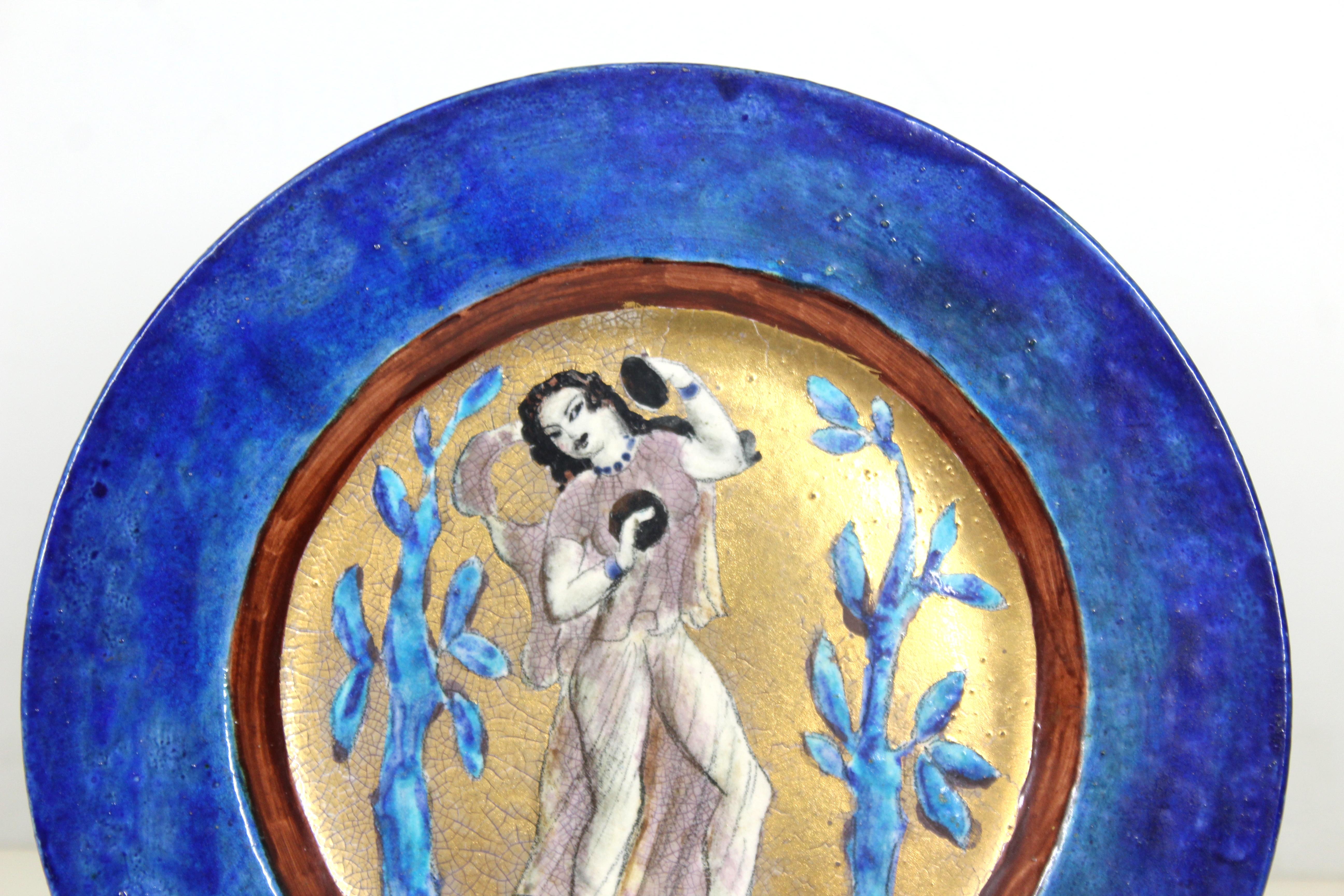 American Art Deco glazed ceramic charger plate in Persian blue and gold, made by Edith Varian Cockcroft (American 1881-1962). The piece presents a hand-painted and gilt center with  a pair of blue trees and an exotic female dancer holding cymbals.