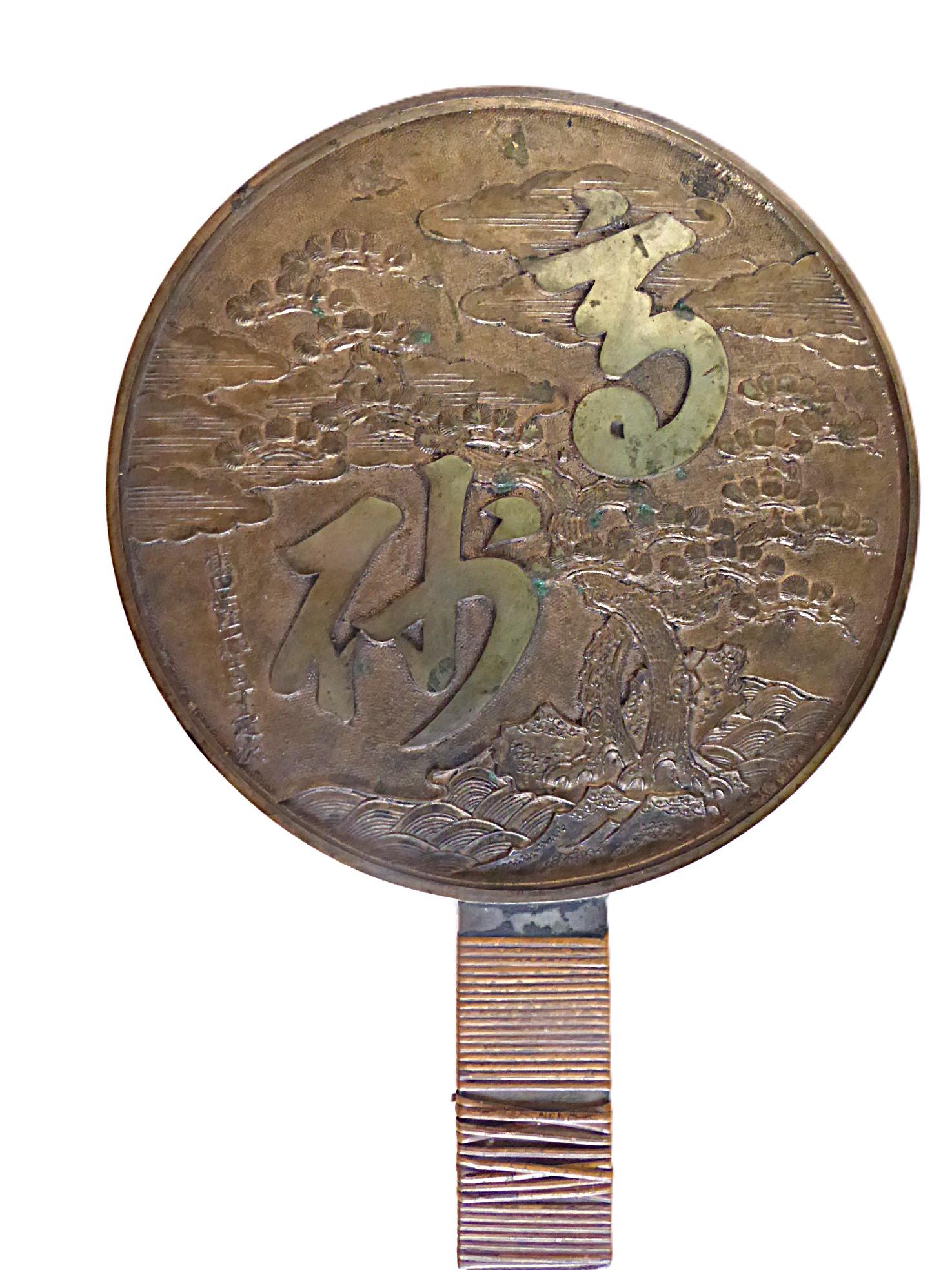 Antiguo espejo japonés de bronce decorado con una escena de nubes arremolinándose sobre dos pinos entrelazados, símbolo de larga vida, en un promontorio con olas rompiendo contra él.  Se superponen grandes caracteres japoneses sobre la escena y la
