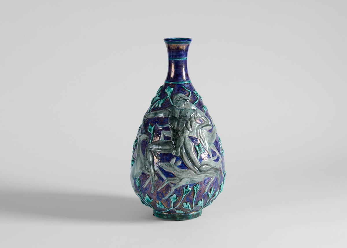Ce remarquable vase en porcelaine date des années 1930 et est l'œuvre de l'un des principaux représentants de la céramique déco française, Edouard Cazaux. Avec cet arrangement irisé de glaçures vertes, bleues et dorées, le céramiste crée une scène