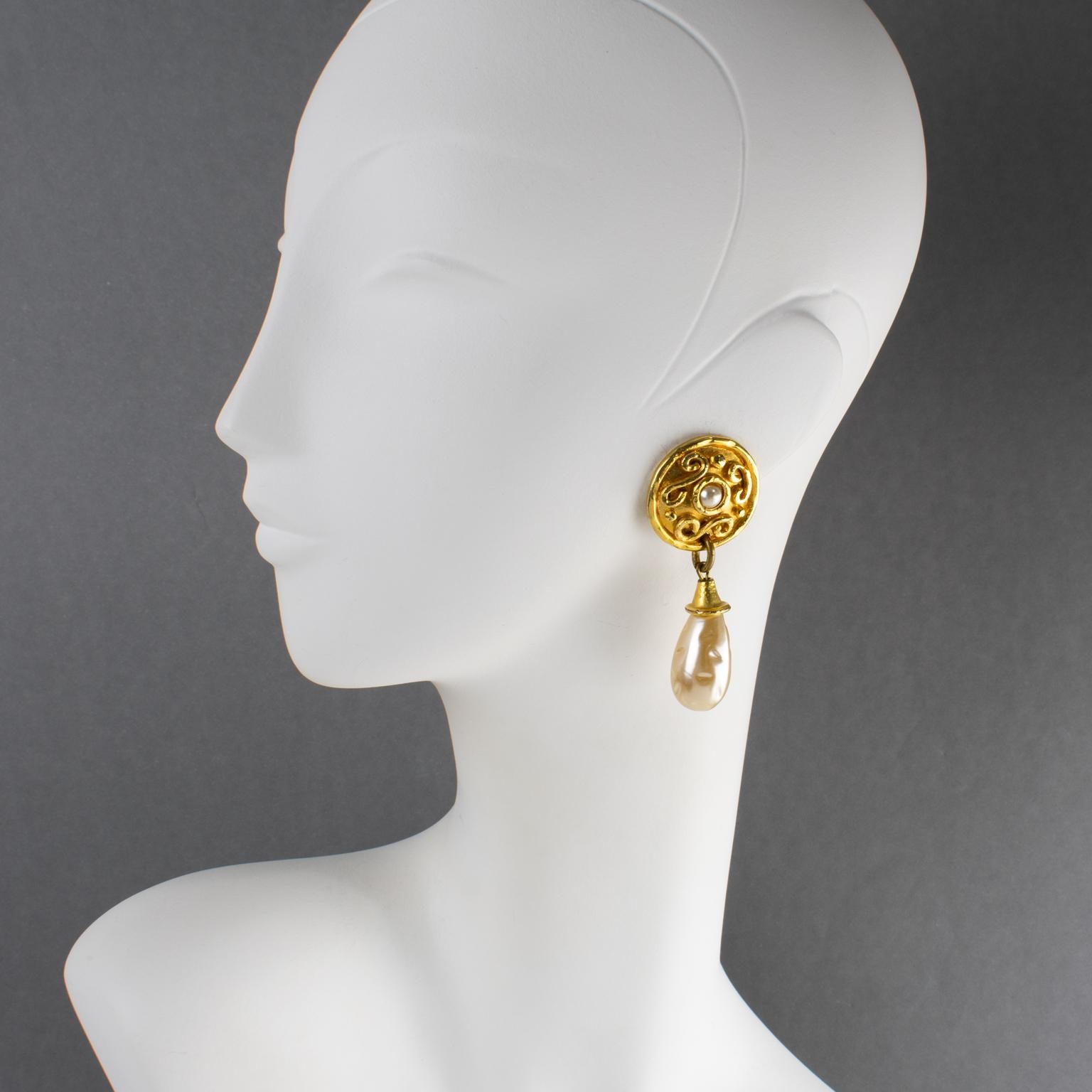 Questi eleganti orecchini a clip hanno un design che ricorda le opere del designer francese Edouard Rambaud. La forma penzolante di ispirazione bizantina è caratterizzata da metallo lucido e dorato, strutturato e ornato, completato da gocce e