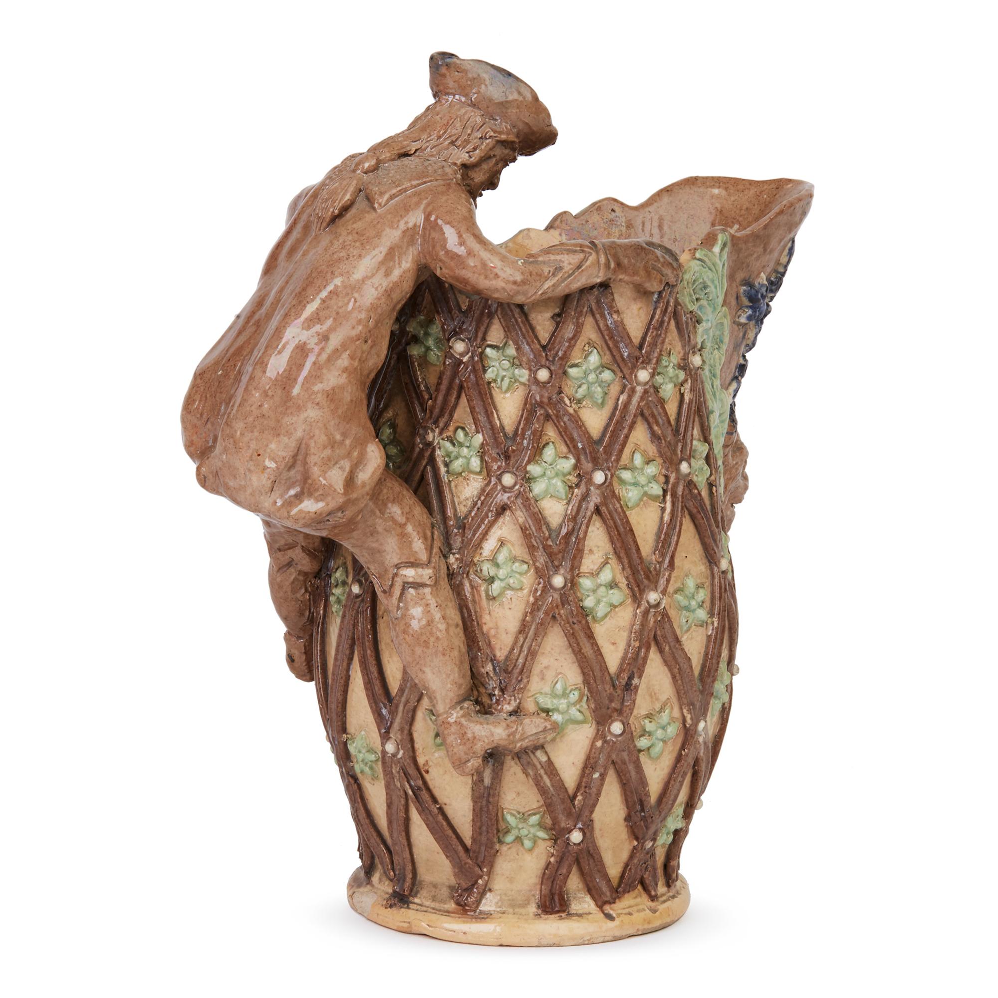 A fine medieval inspired Castle Hedingham art pottery jug by Edward Bingham and dating from the latter 19th century. The jug is hand modeled with a cavalier like figure forming the handle perched on the side of the jug and peering down into the jug.