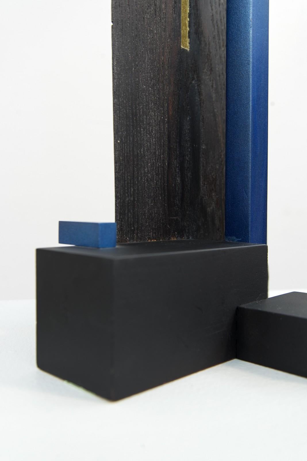 A single vertical shard of milled timber in charred black and pierced at the centre by a narrow vertical window, tapers upwards from a base constructed of painted wood orthogonals. A slender wood rectangle in sapphire blue rises parallel to the
