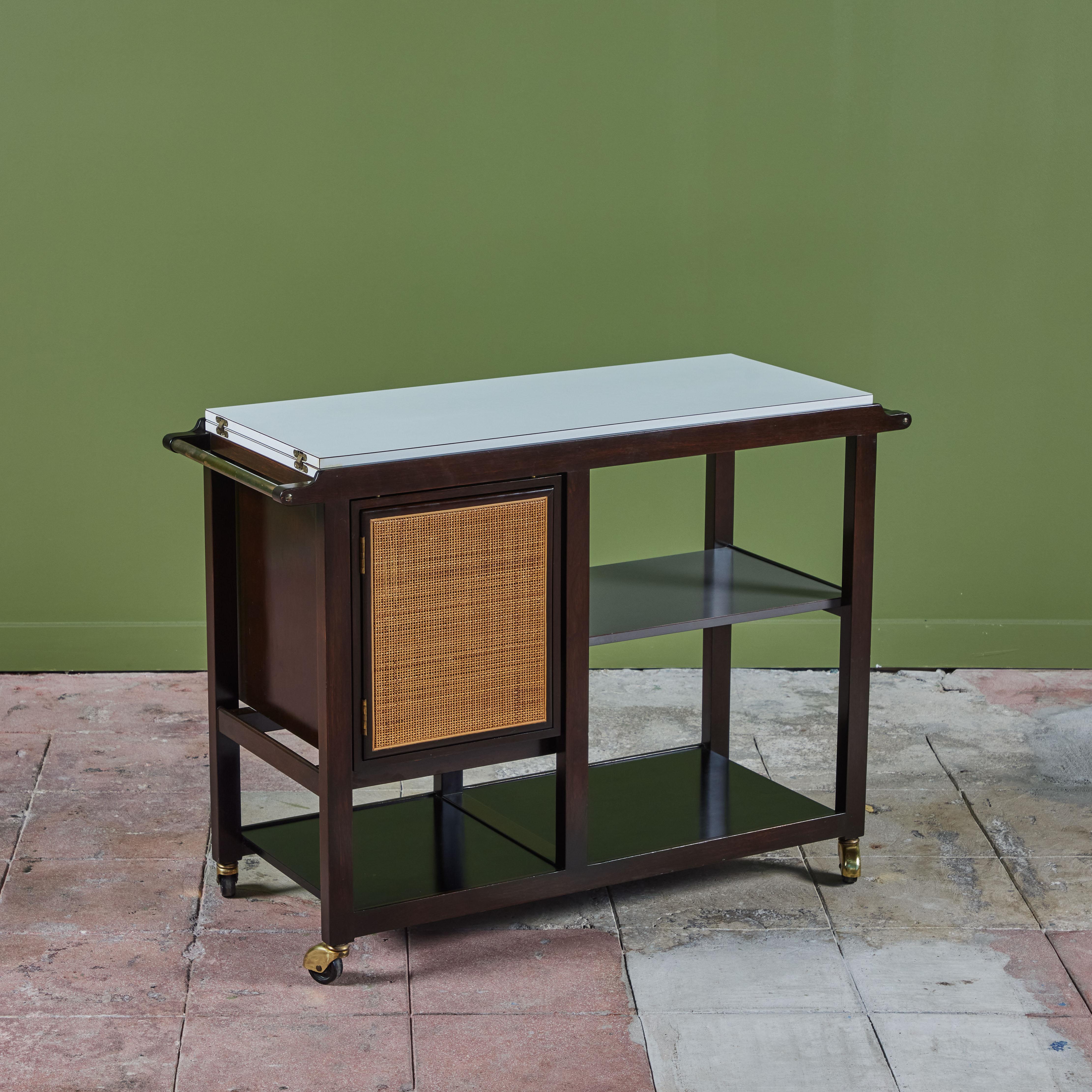 An ebonized bar cart by Edward Wormley for Dunbar, USA, c.1960s. The cart features a white laminate flip top surface with a double sided caned door for storage. The cart has three additional shelves and rests on brass casters for easy mobility.