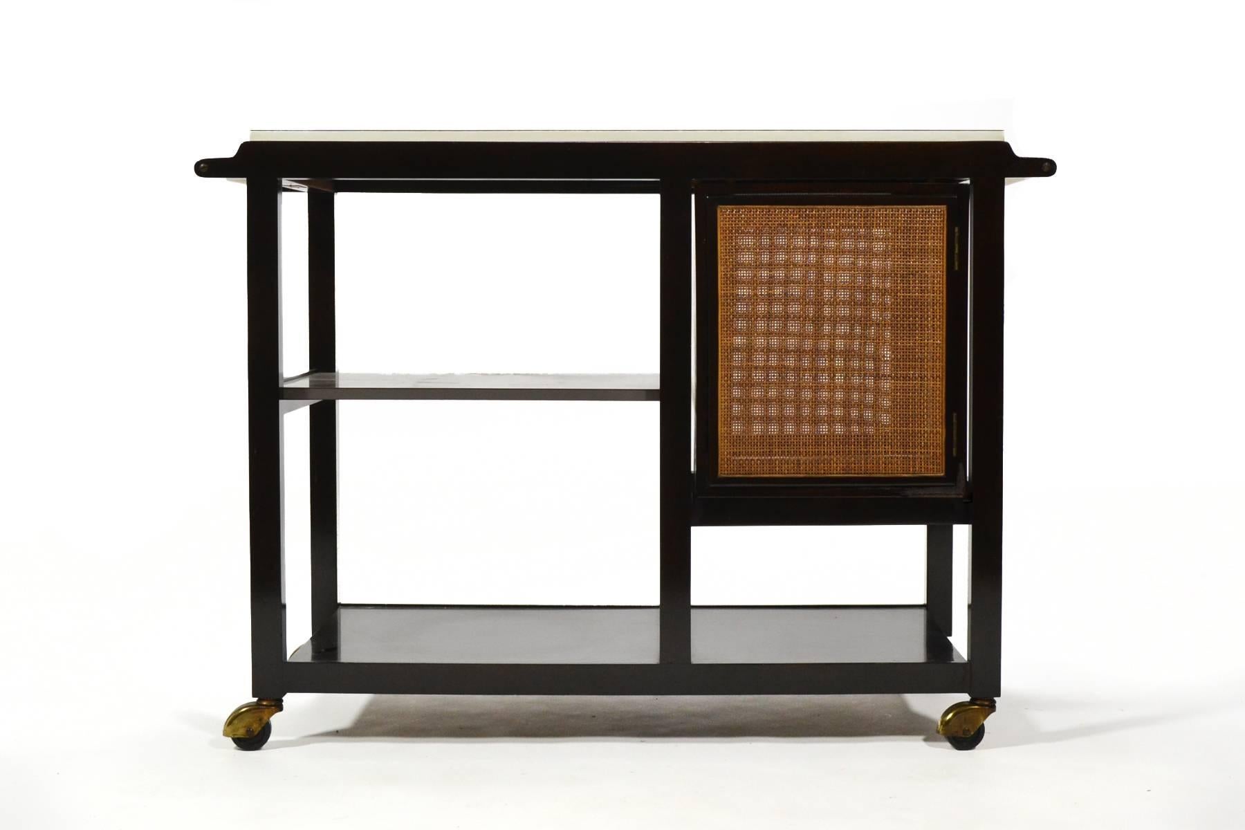 This beautifully designed and detailed rolling cart by Edward Wormley has a lovely arrangement of shelves, a cabinet with cane front doors and a highly functional mica top that flips open to double in size. Appointed with two brass handles and