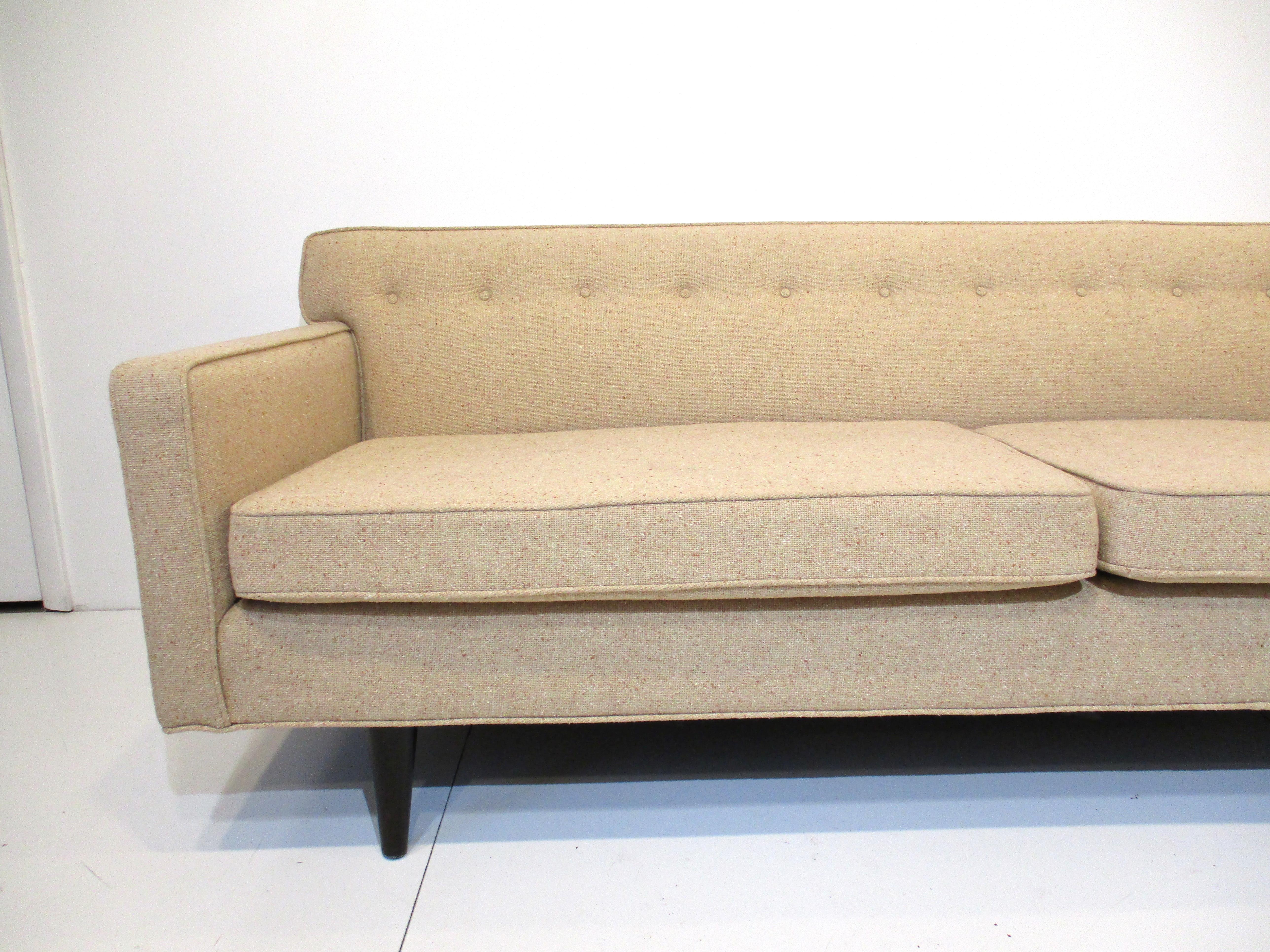 A low profile sofa in an oatmeal toned salt and pepper styled fabric with dark ebony toned mahogany tapered legs . Very well crafted having two loose lower seat cushions and buttons to the backside, designed by Edward Wormley for the Dunbar