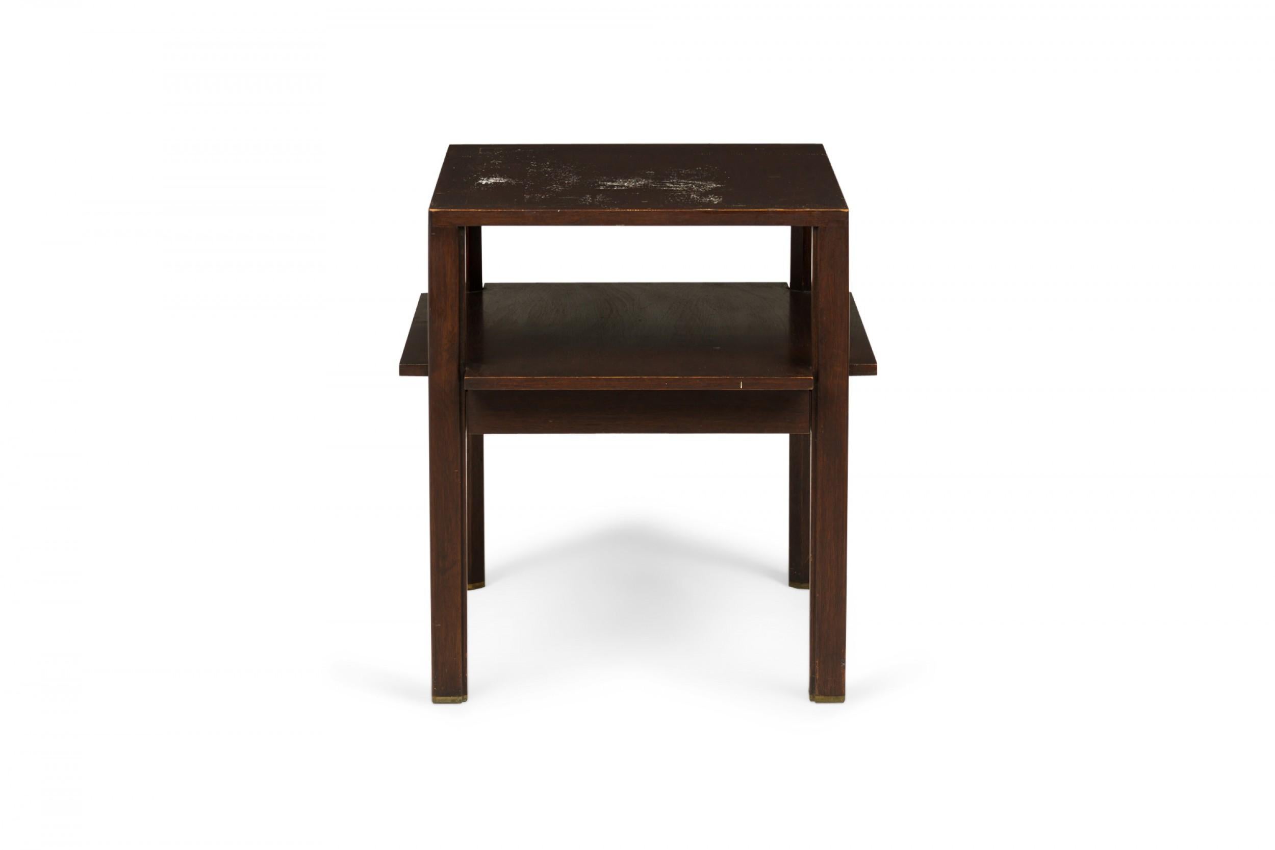 American Mid-Century wooden two-tier end / side table with a dark finish, square top, and slightly larger lower stretcher shelf above a shallow single drawer, supported by four square legs with rounded corners, ending in brass sabots. (EDWARD